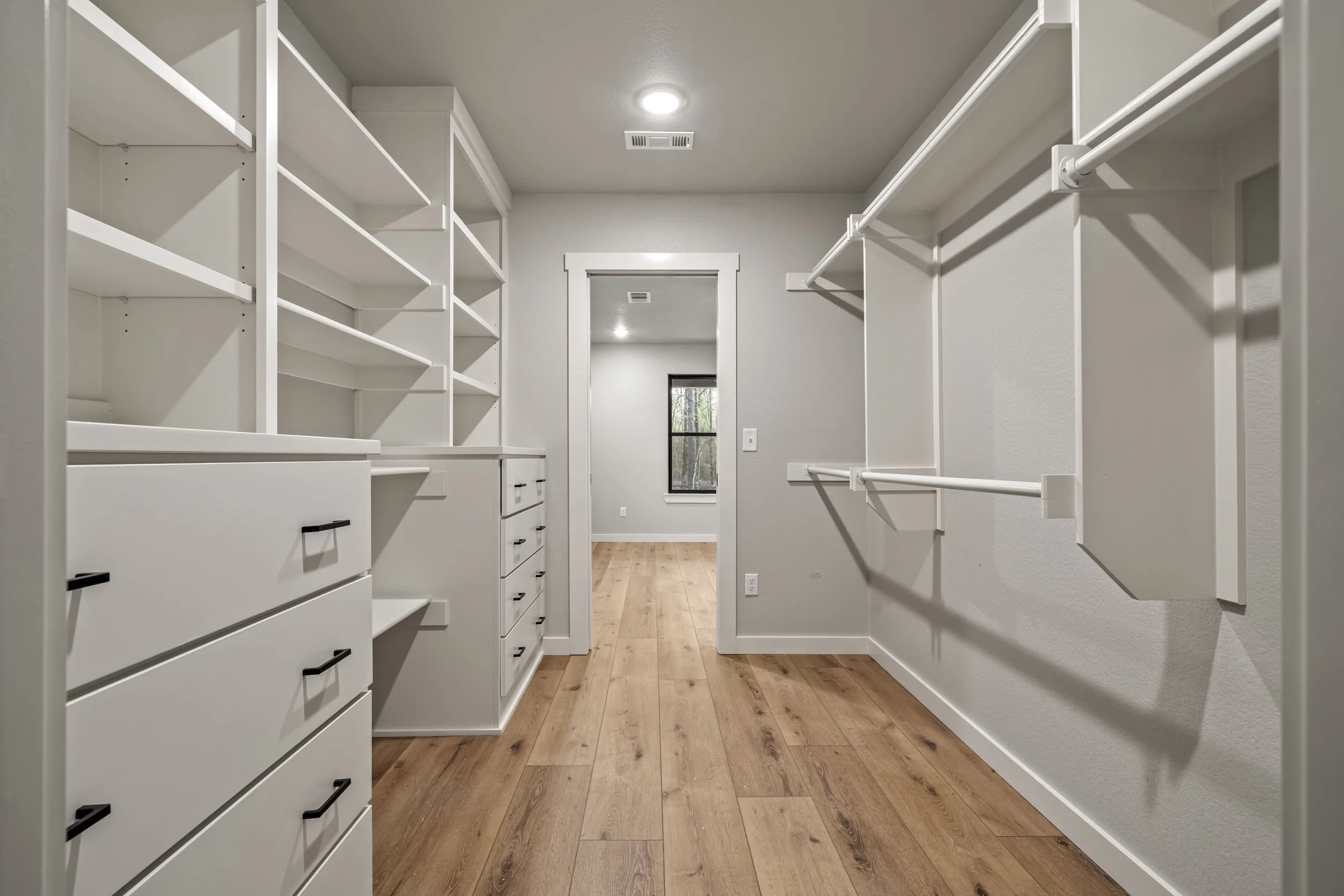 Empty white walk-in closet with built-in shelves, drawers, and hanging rods, featuring wooden flooring and a window at the far end.