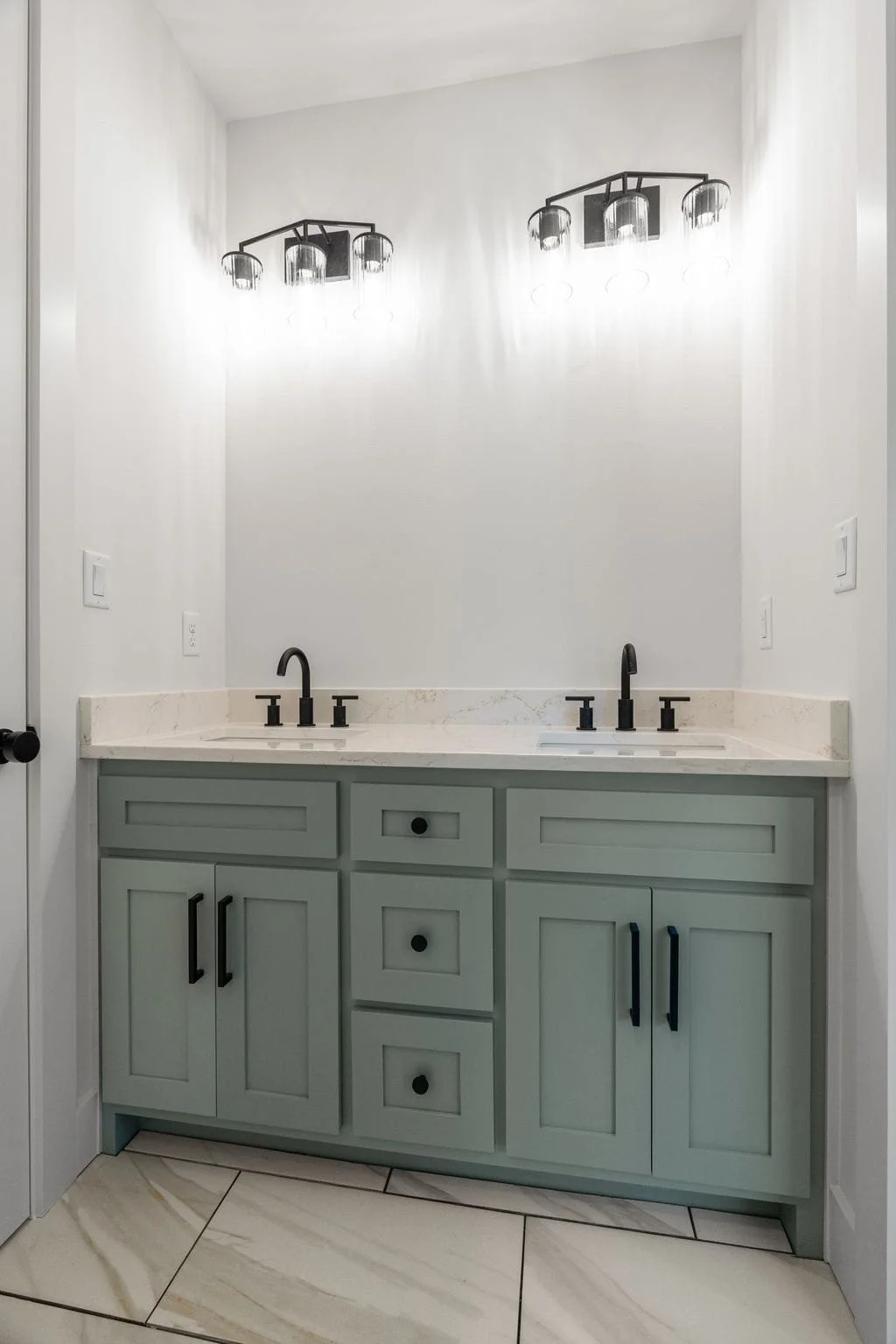 Modern bathroom vanity with double sinks, black faucets, light green cabinets, white marble countertop, and black light fixture overhead.
