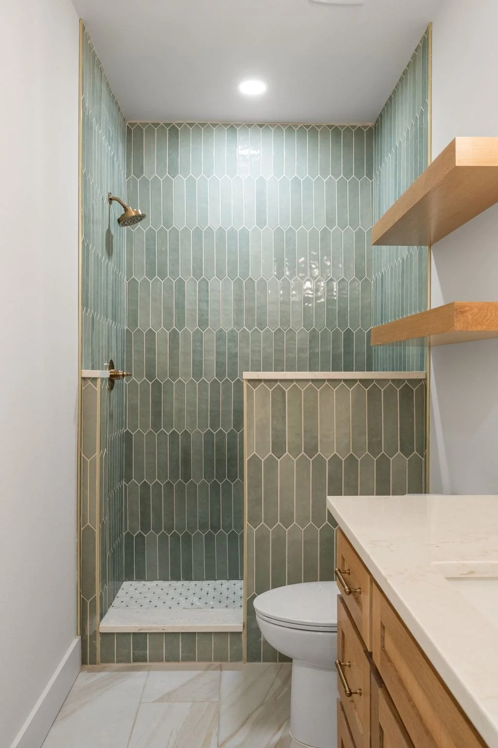 Small bathroom with a green tiled shower, a toilet, wooden shelves, and a wooden vanity with a white countertop.
