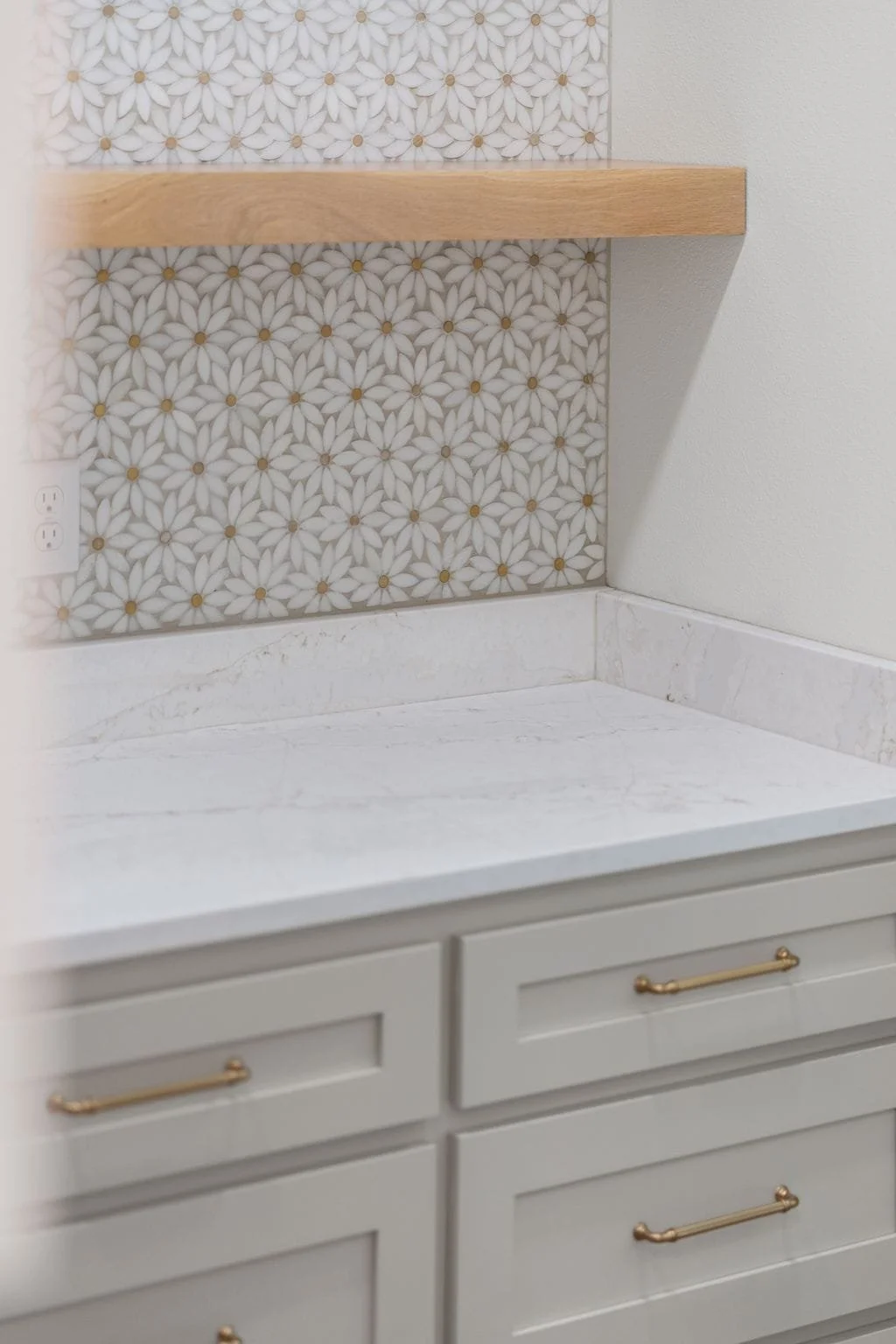 Bathroom with white cabinetry, a marble countertop, a decorative tile backsplash with a floral pattern, a wooden shelf, and an electrical outlet.