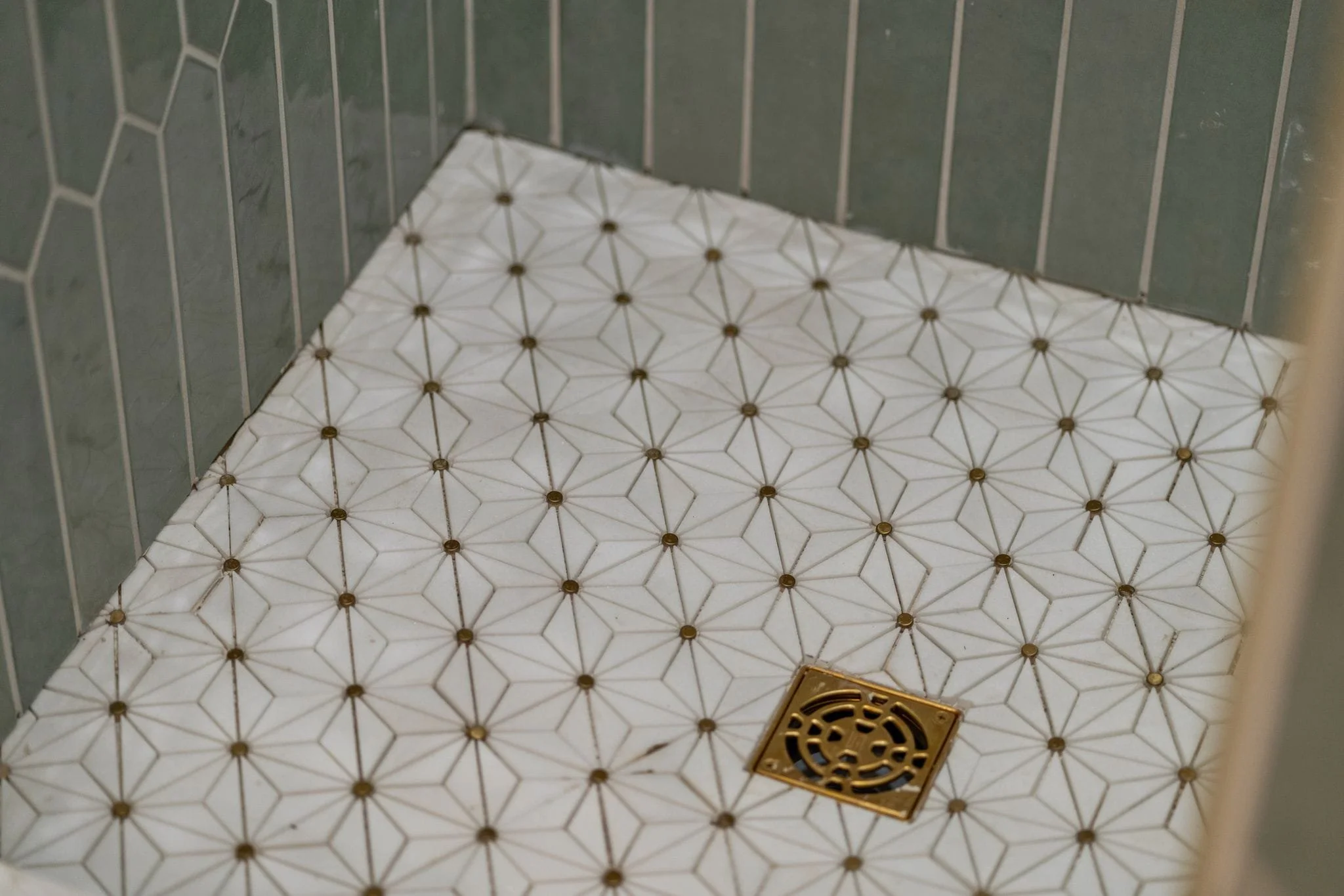Close-up of a corner of a bathroom floor with white tiles featuring a geometric pattern and brown accents, along with a drain cover.