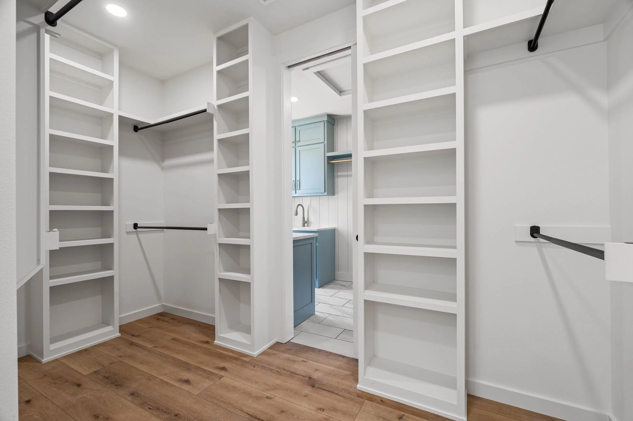 Empty walk-in closet with white shelving, hanging rods, and a view into a laundry or utility room with blue cabinets and a sink.