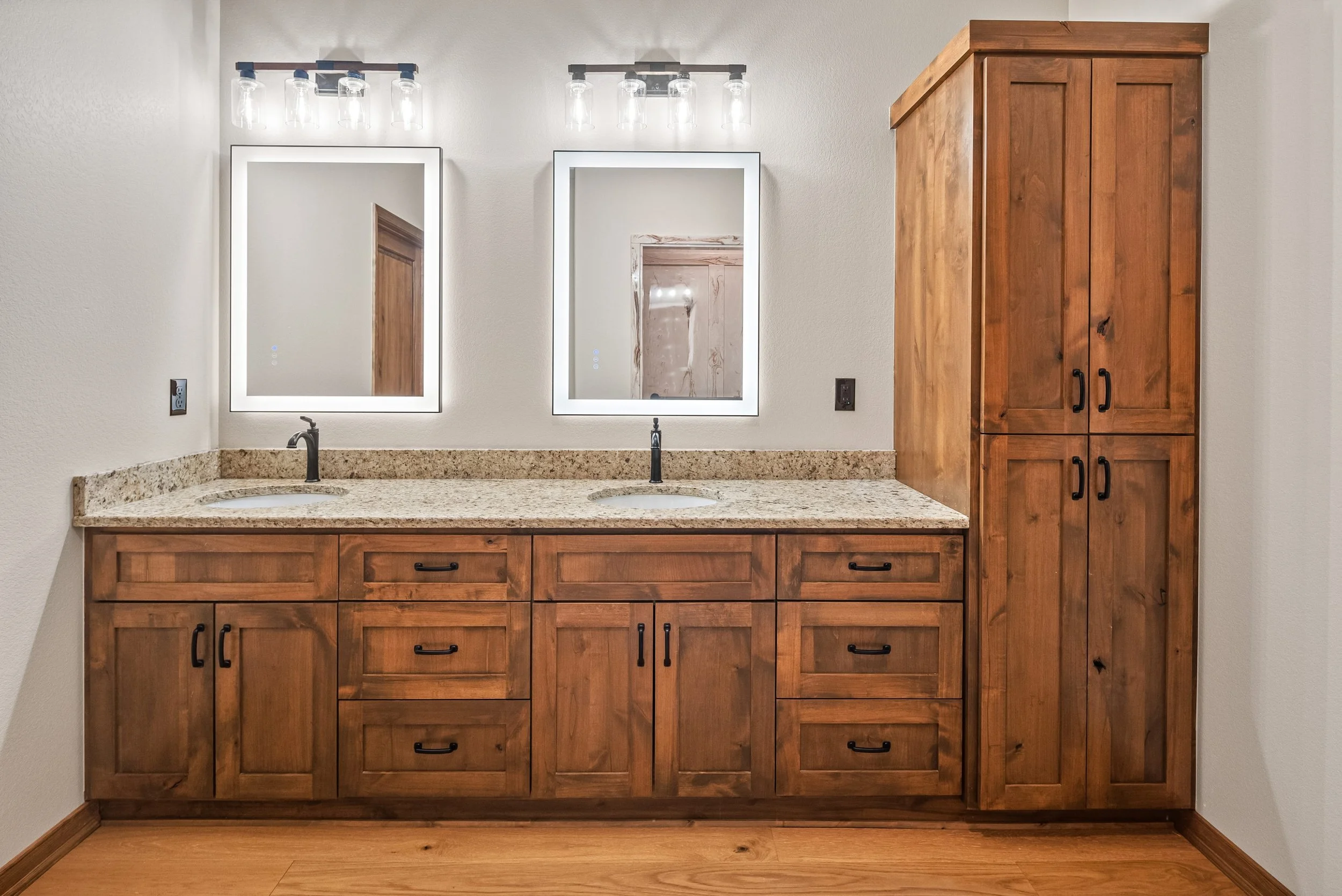 Bathroom vanity with two sinks, granite countertop, wooden cabinets, two illuminated mirrors, and a tall matching wooden cabinet to the right.