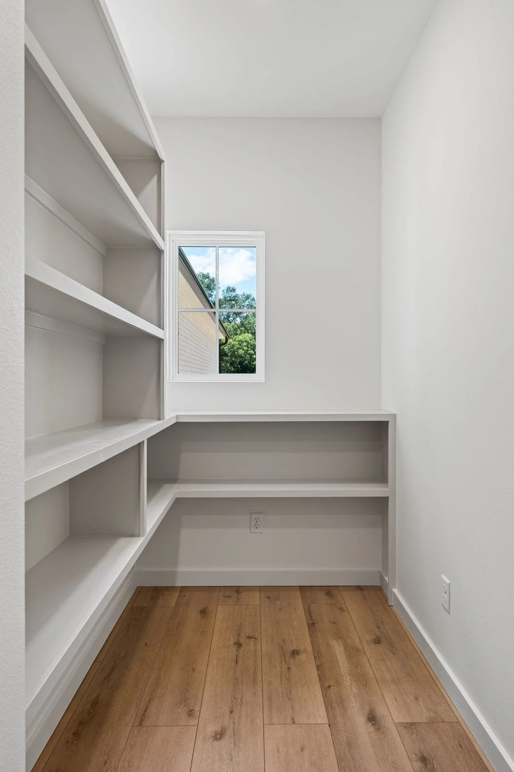 Empty small room with white walls, wooden floor, built-in white shelves, and a small window showing trees outside.