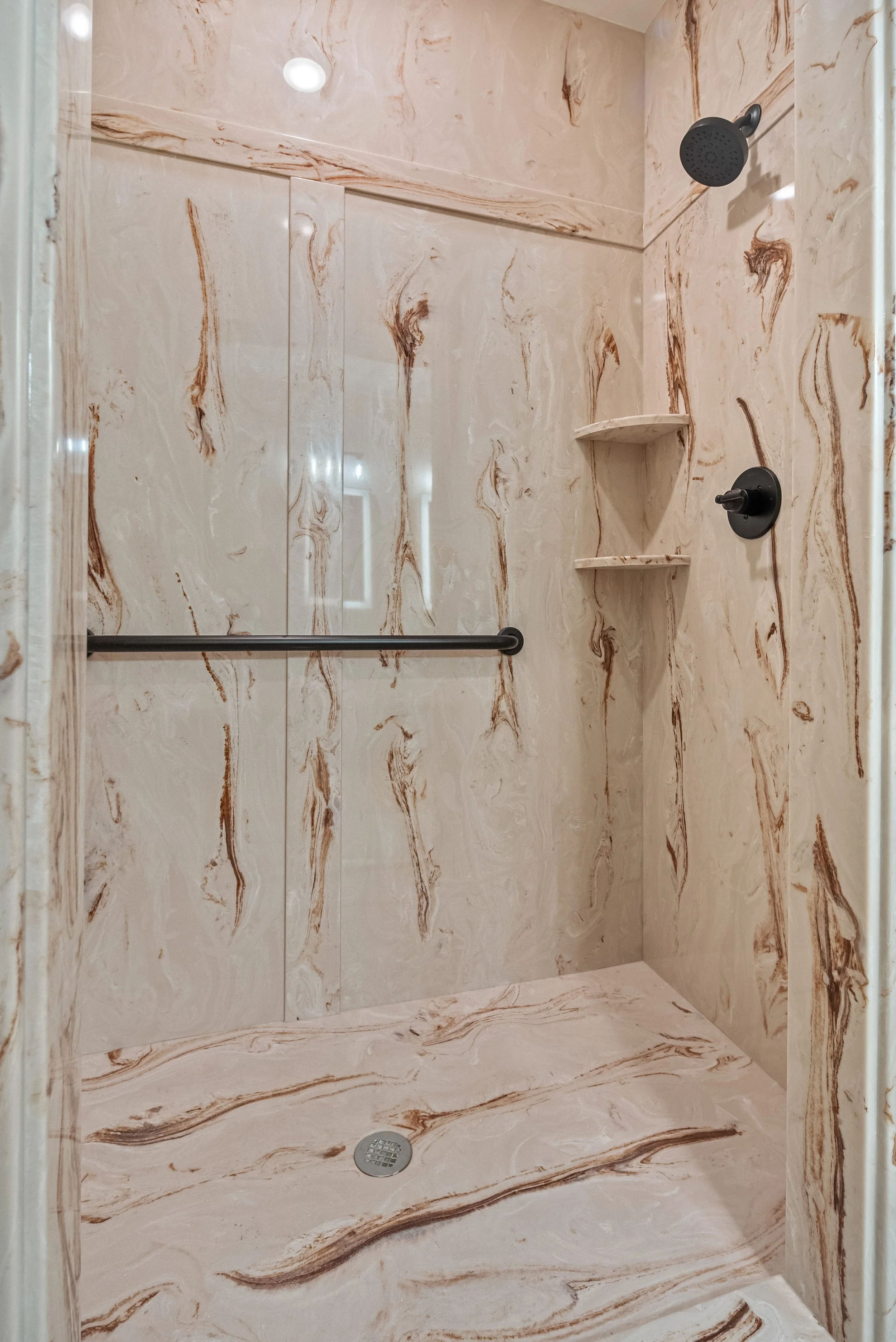 A shower with beige marble walls and floor, a black showerhead, a black shower control, a black grab bar, and built-in corner shelves.