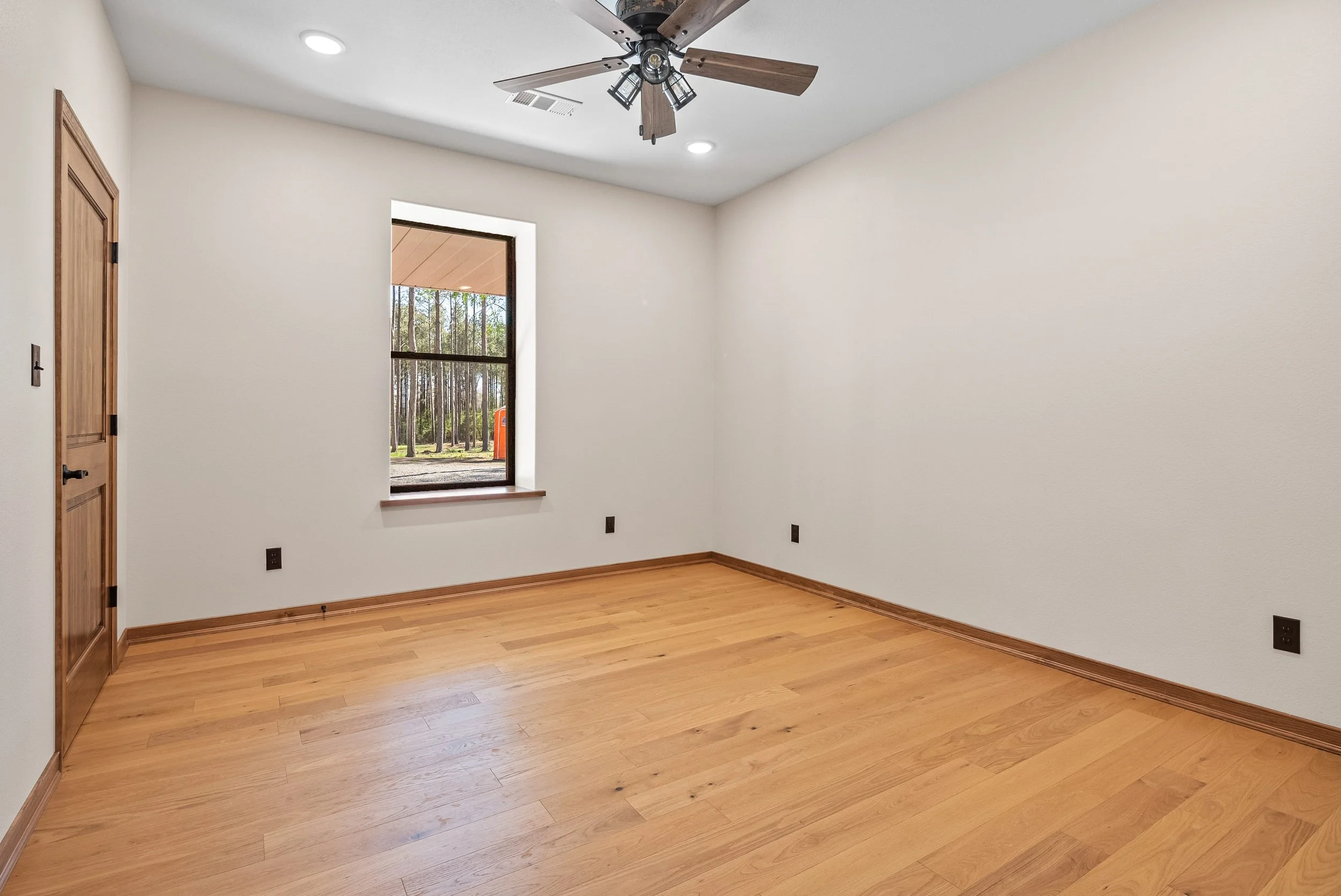 Empty room with hardwood floor, white walls, a window showing outdoor trees, a ceiling fan, and recessed lighting.