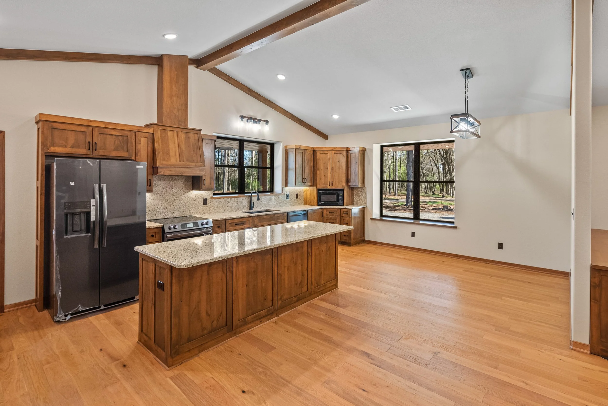 Kitchen with wooden cabinets, granite countertops, stainless steel refrigerator, microwave, daylight windows, hardwood flooring, and a modern hanging light fixture.