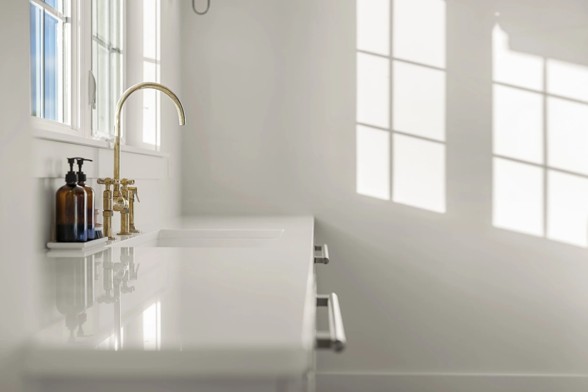 White kitchen sink with gold faucet and soap bottles on white countertop, bright sunlight streaming through large windows.