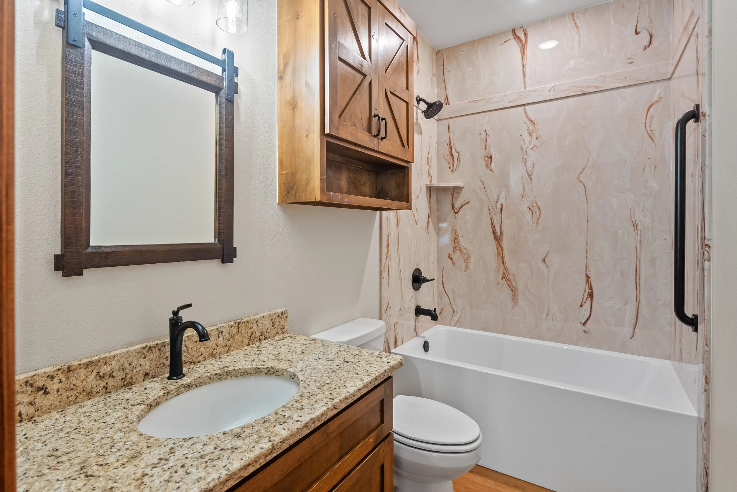 A bathroom with a granite countertop sink, a toilet, a wood cabinet, a mirror, and a walk-in shower with marble walls and black fixtures.
