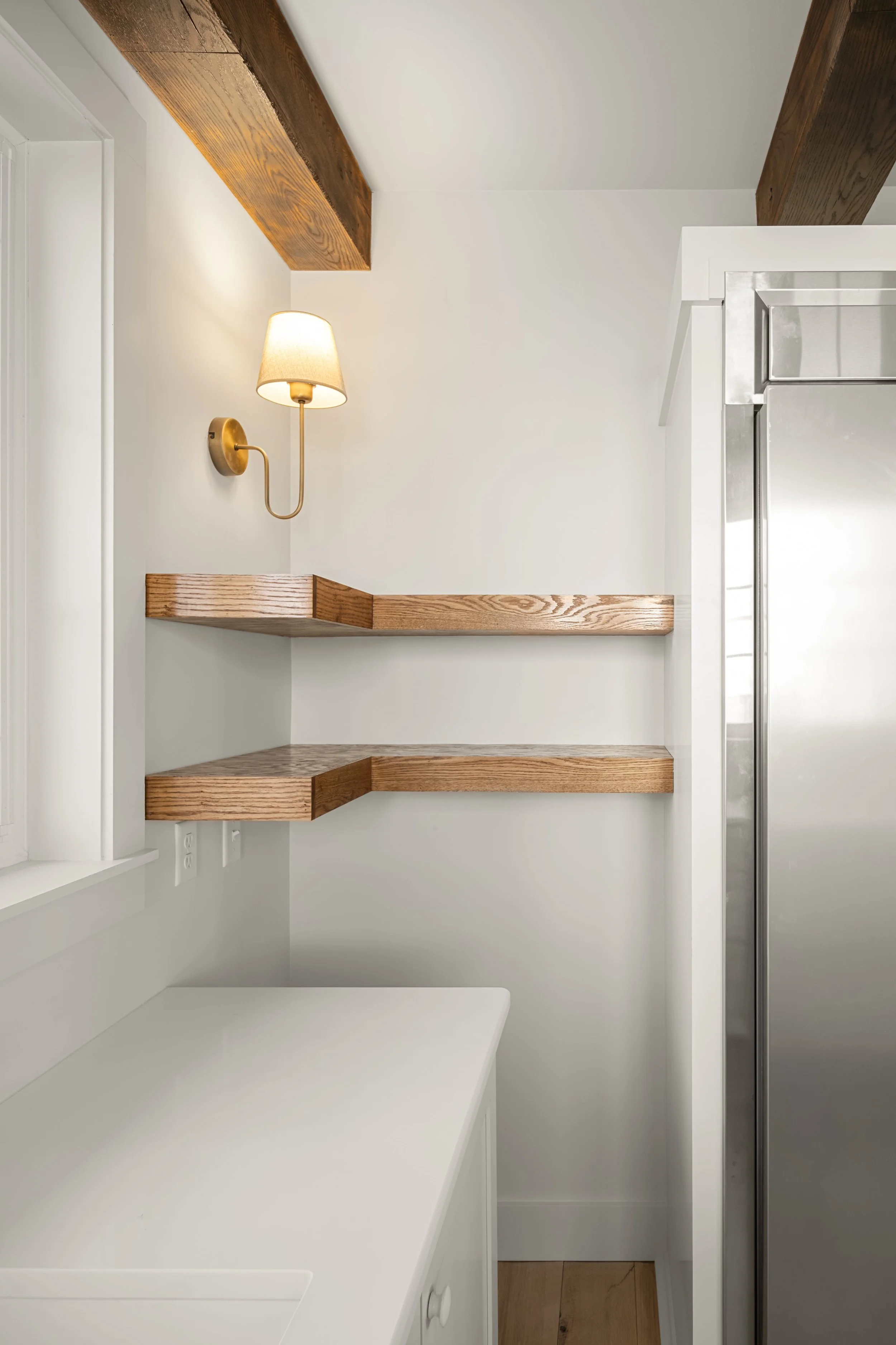 Interior of a kitchen corner featuring white cabinets, exposed wooden beams, two wooden shelves, a wall-mounted lamp, and part of a stainless steel refrigerator.