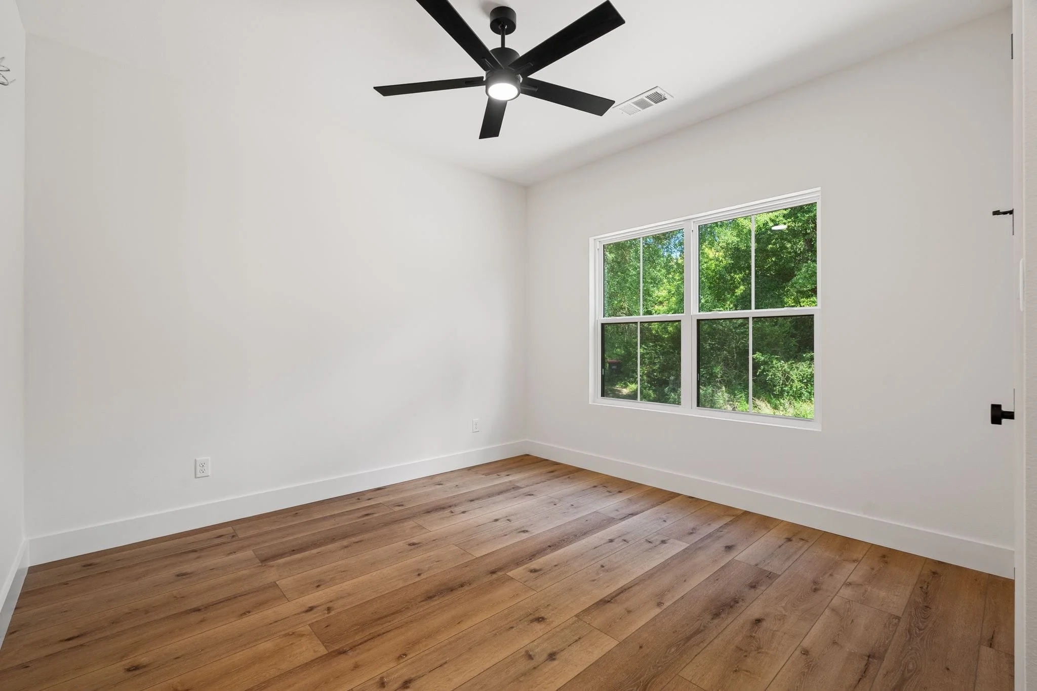 Empty bedroom with white walls, a window showing trees outside, a ceiling fan with lights, and hardwood flooring.