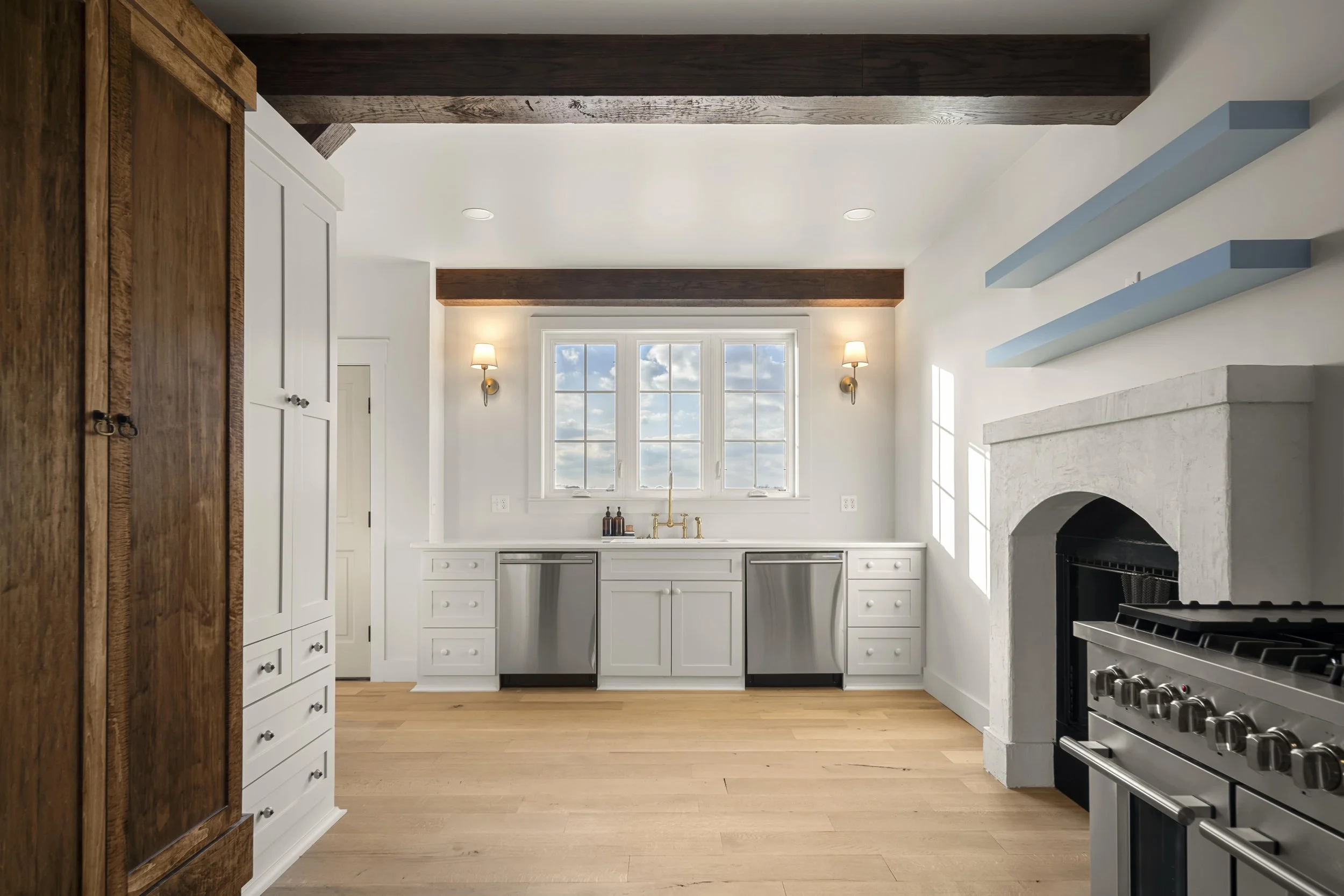 Bright kitchen with white cabinets, stainless steel dishwasher, and a large window with a view of the cloudy sky. Wooden beams on the ceiling, a fireplace on the right, and light wooden flooring.
