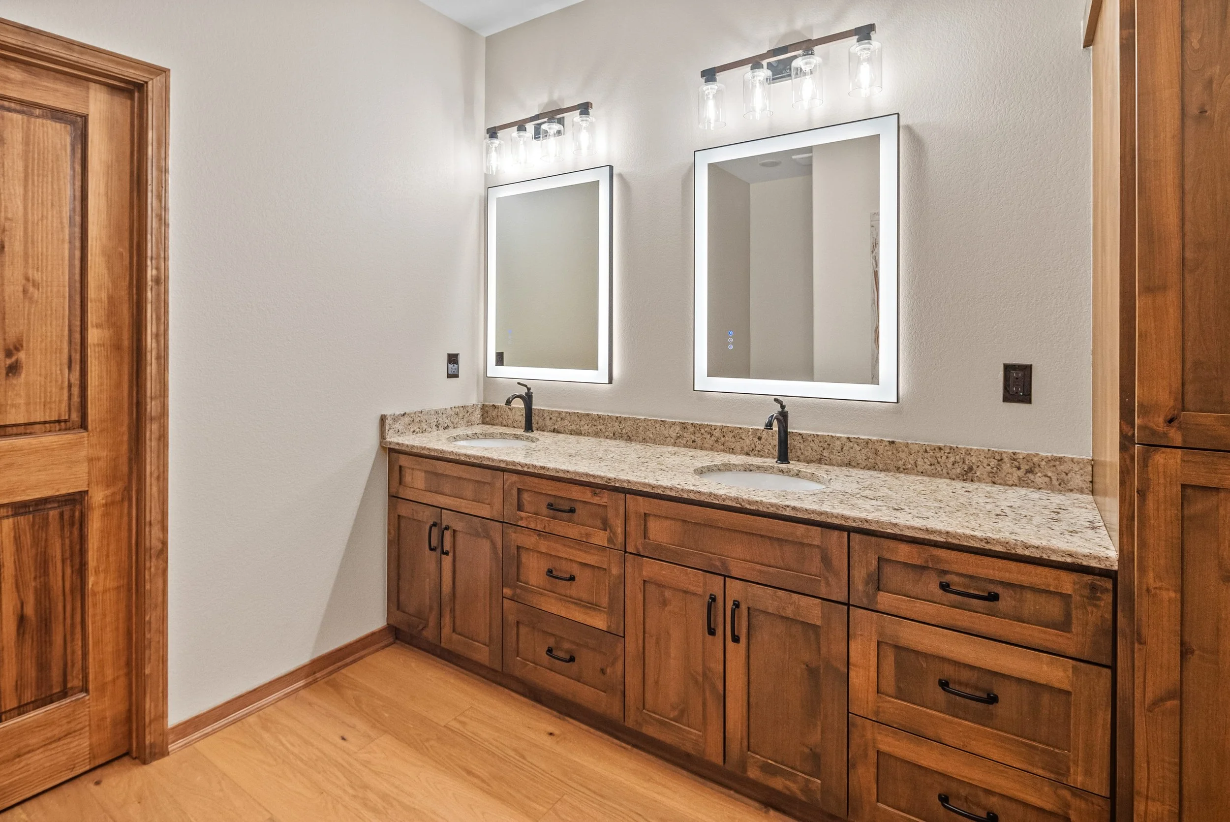Double vanity bathroom with two mirrors, wooden cabinets, granite countertop, and black faucets, illuminated by modern light fixtures.