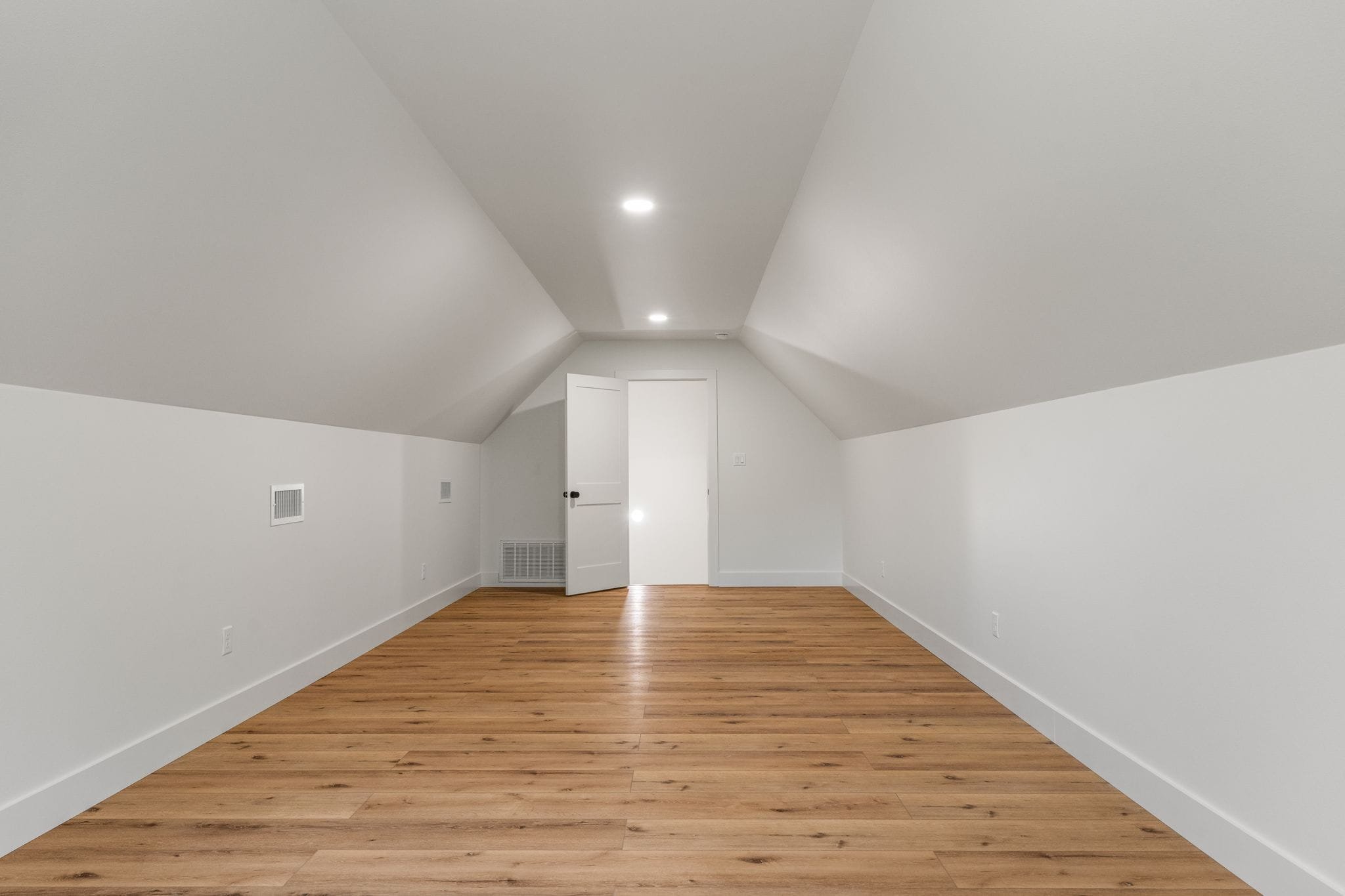 Empty room with white sloped ceiling, white walls, wooden flooring, and a door.
