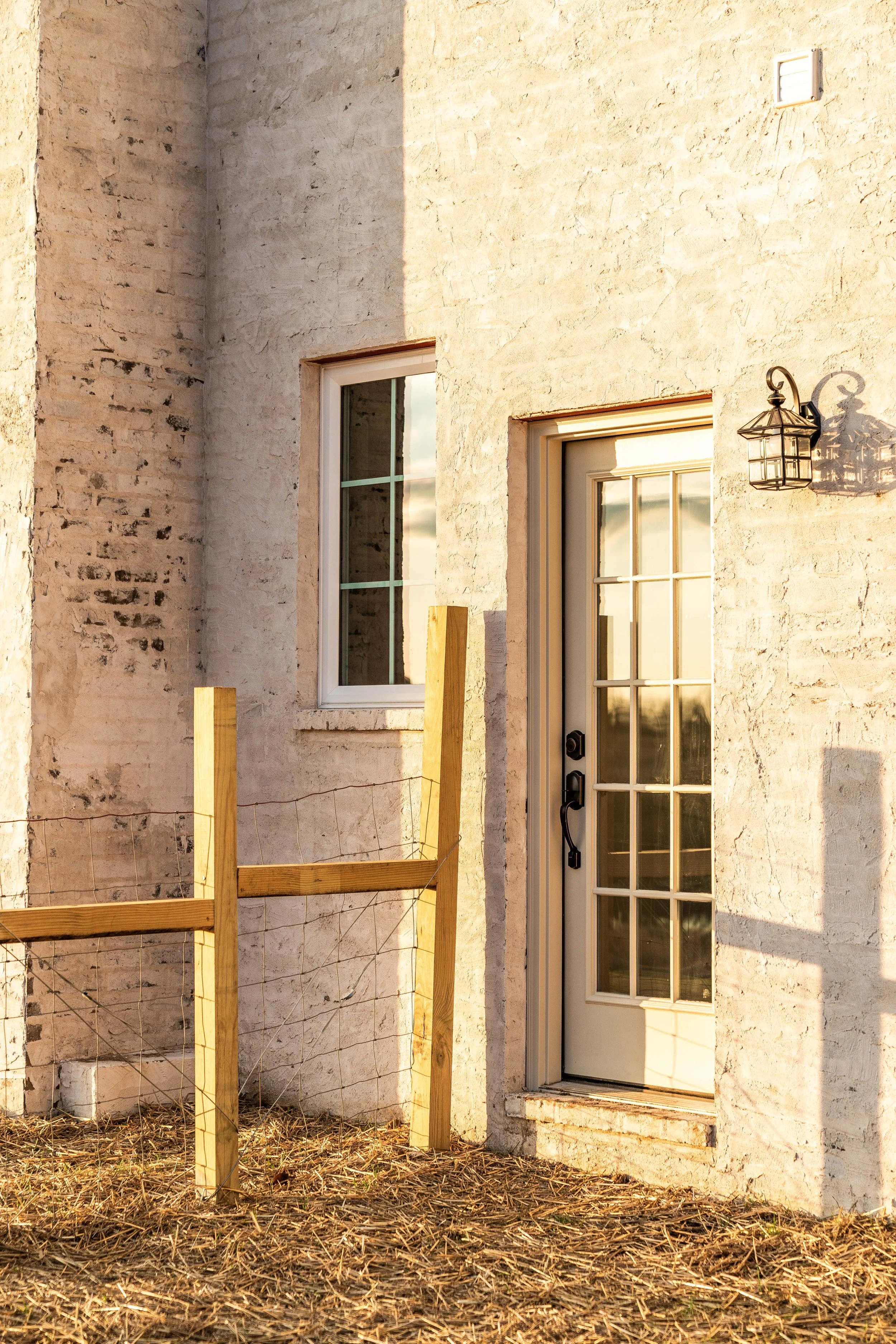 Close-up of a building's exterior wall with a window and door, and a small outdoor light fixture, photographed in warm sunlight.
