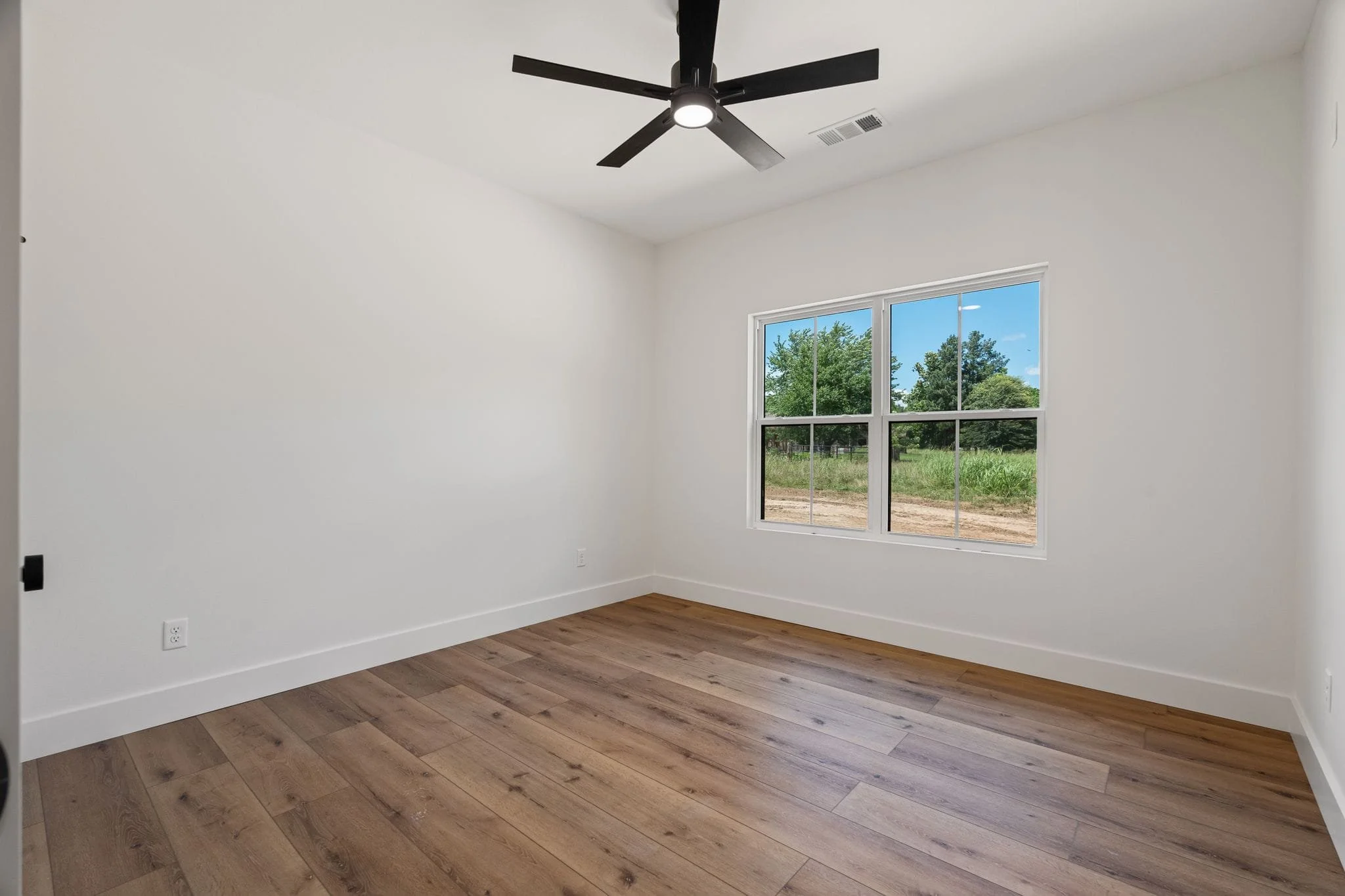 Empty room with white walls, hardwood floors, a large window showing green trees and blue sky, a black ceiling fan, and electrical outlets.