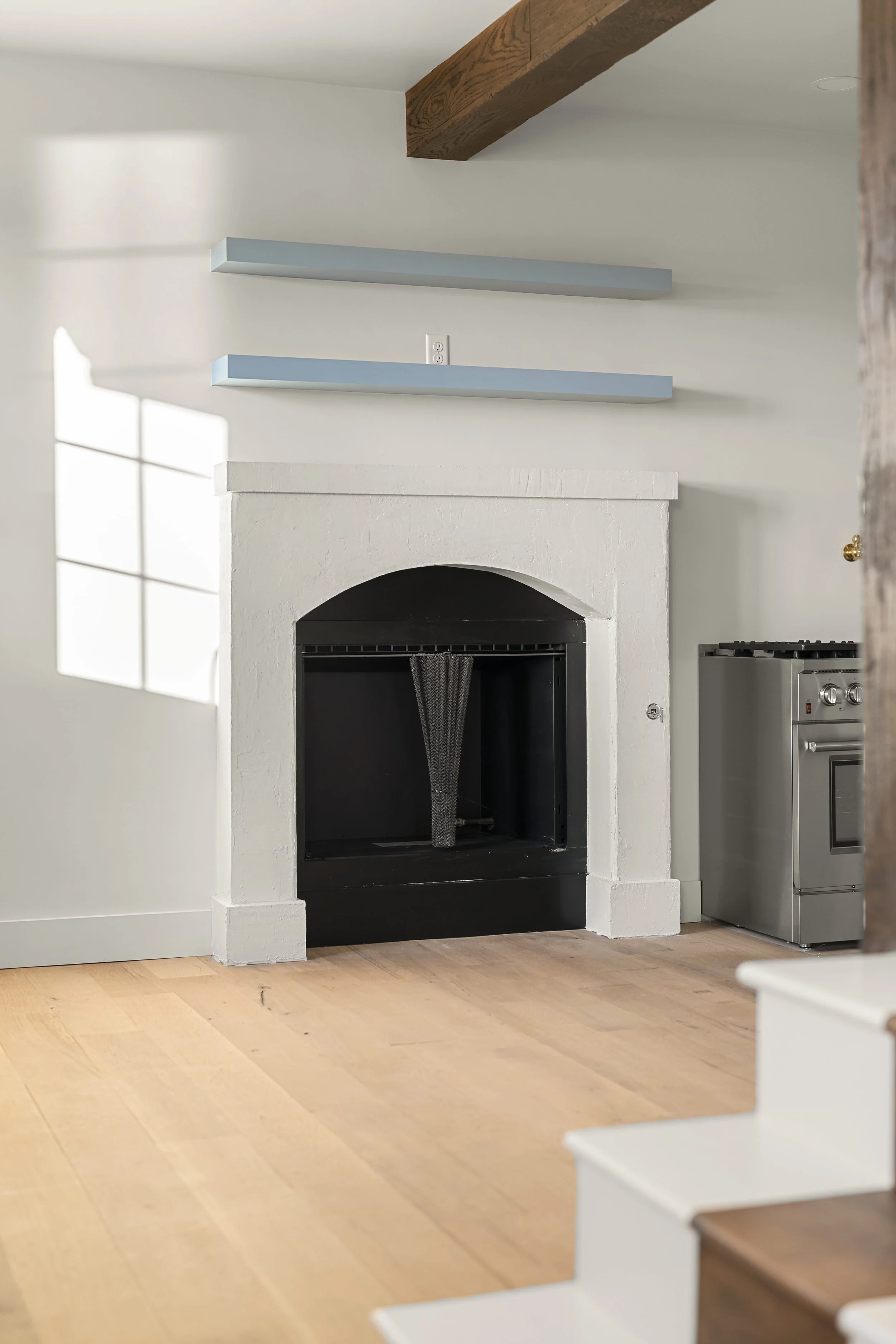 Interior of a modern living space with a white fireplace, light wood floors, and a window casting sunlight on the wall. There are two light blue wall-mounted shelves above the fireplace, and a stainless steel stove is visible on the right.