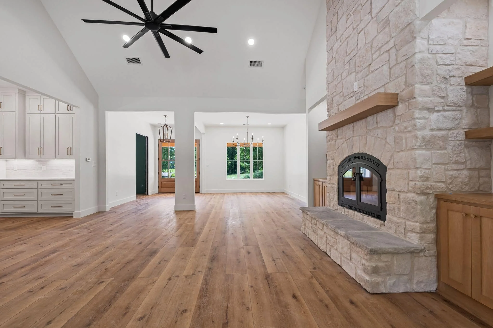 Interior view of a modern room with white walls, hardwood flooring, a stone fireplace on the right, large windows in the background, and a black ceiling fan with lights.
