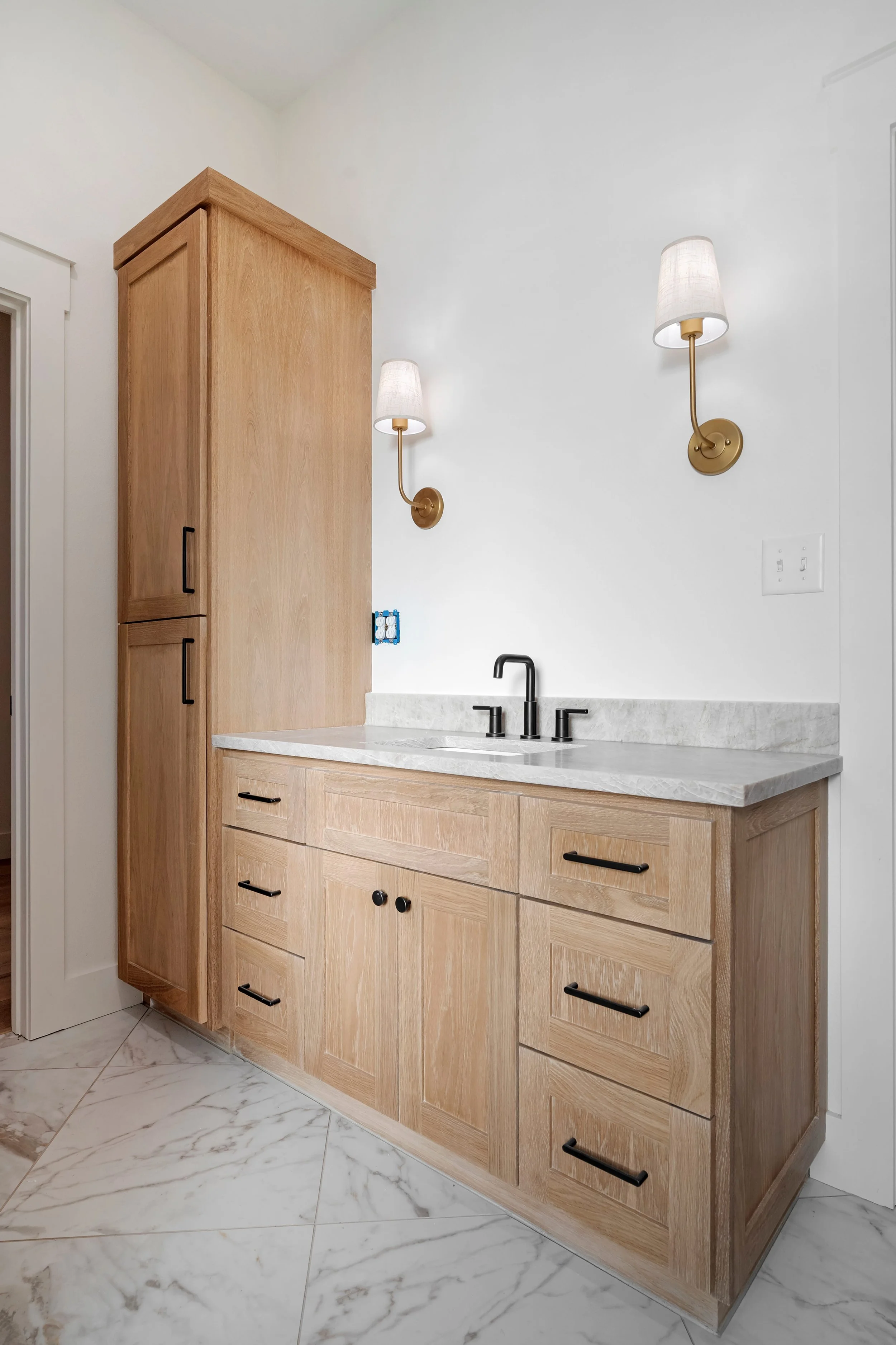 A modern bathroom vanity with a light wood cabinet, a marble countertop, a black faucet, and black handles. Two wall-mounted lamps with white shades on a white wall, and white marble tile flooring.