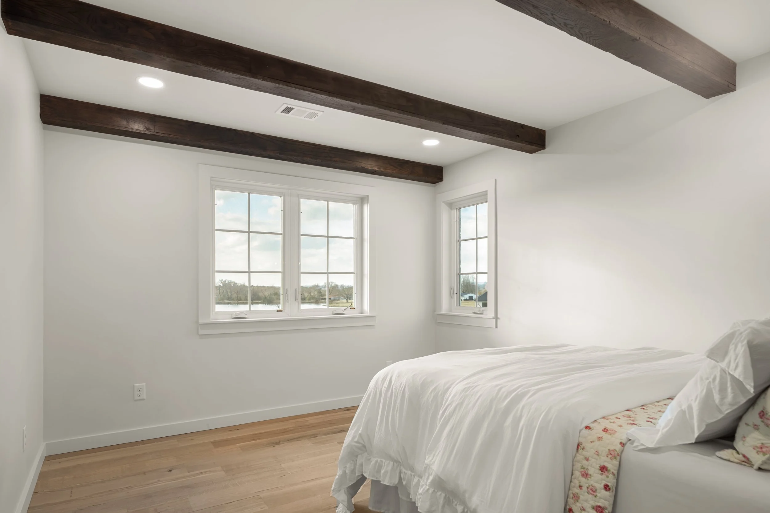 A bedroom with white walls, wooden beams on the ceiling, hardwood floor, and a bed with white bedding near three windows showing a landscape view.