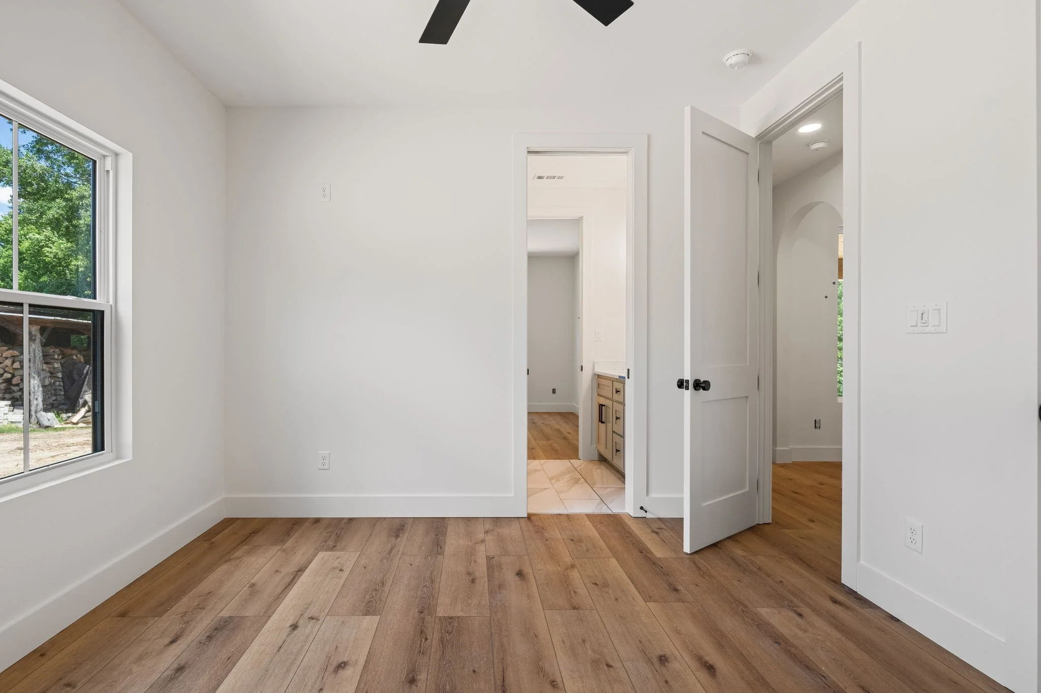 Interior view of a room with white walls, hardwood floors, a window showing greenery outside, and open doorways leading to other rooms.