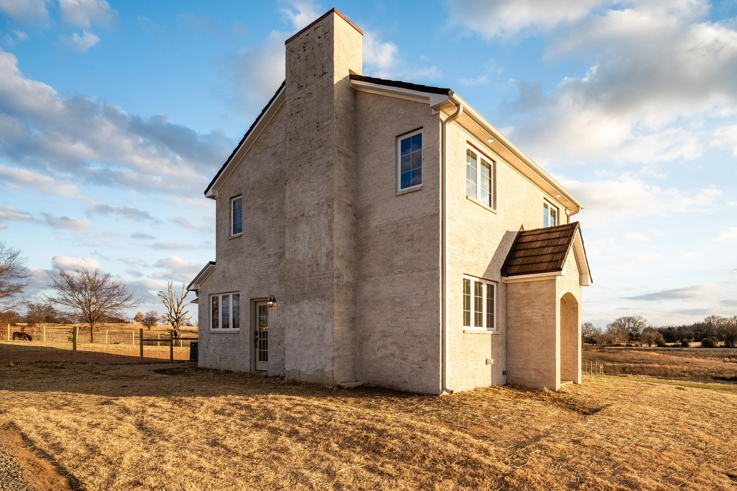 A two-story house with a beige brick exterior, multiple windows, and a small porch with an arched entryway, set against a partly cloudy sky and a rural landscape.