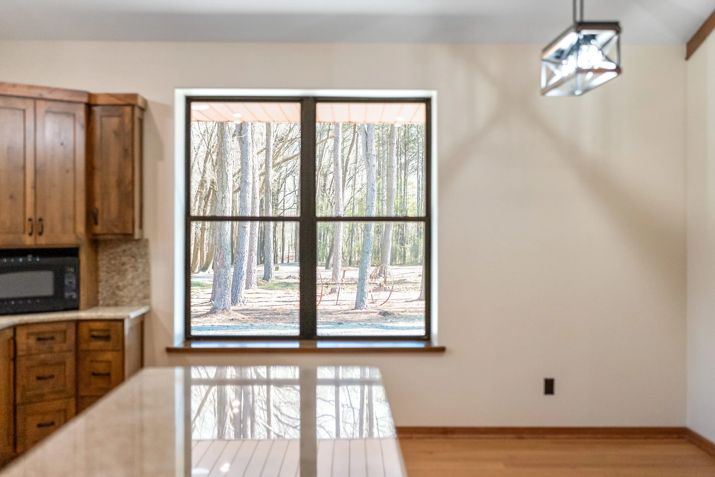 Interior of a kitchen with a large window showing trees outside, wooden cabinets on the left, hanging light fixture, and a counter.