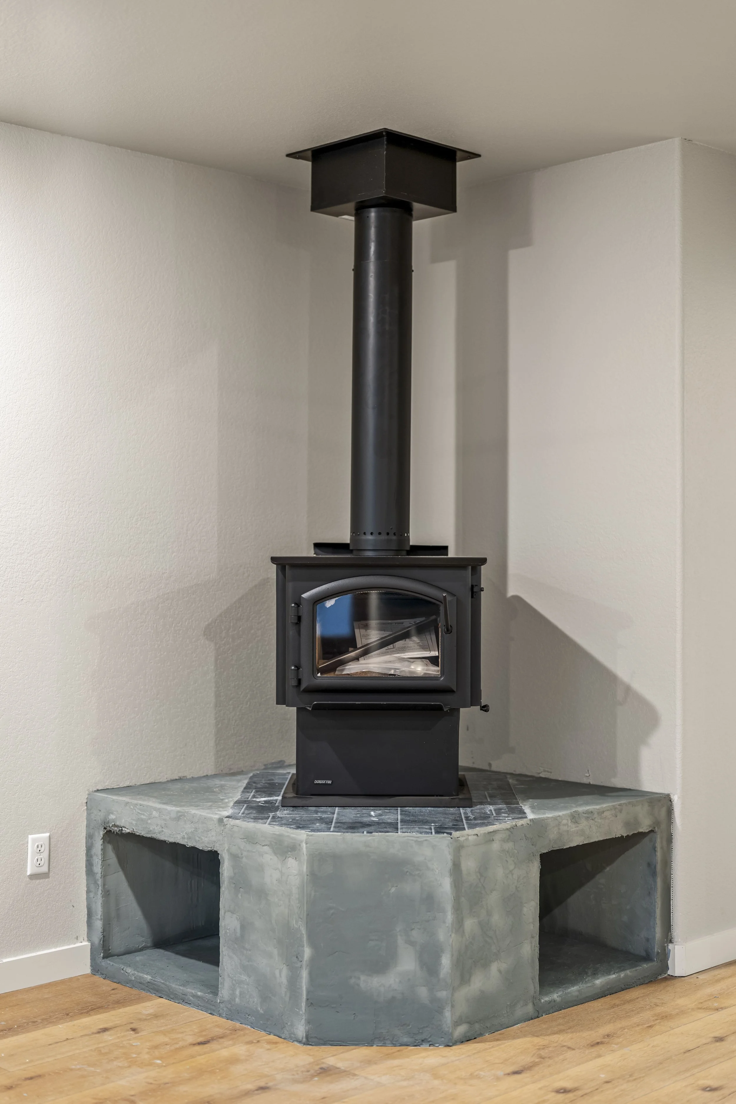 A black wood stove with a glass door, sitting on a concrete hearth with two open compartments underneath, in a room with white walls and wooden floor.