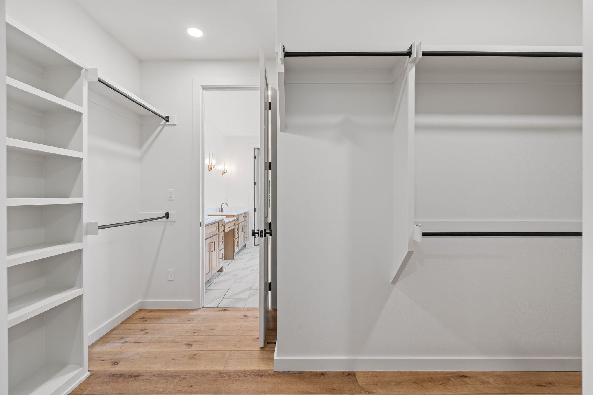 Empty walk-in closet with white built-in shelves and black hanging rods, view towards bathroom with vanity and sink.