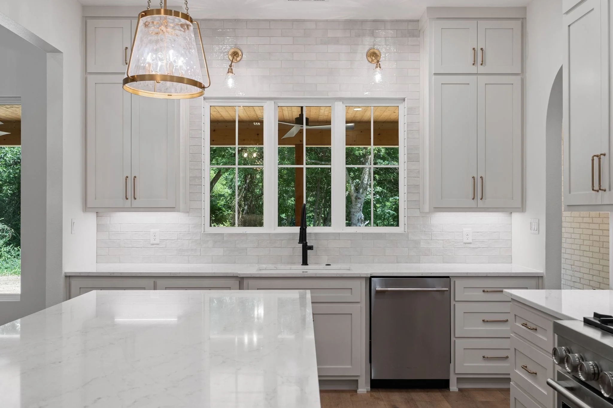 Modern white kitchen with large window, white cabinets with gold handles, marble countertops, and a black faucet.