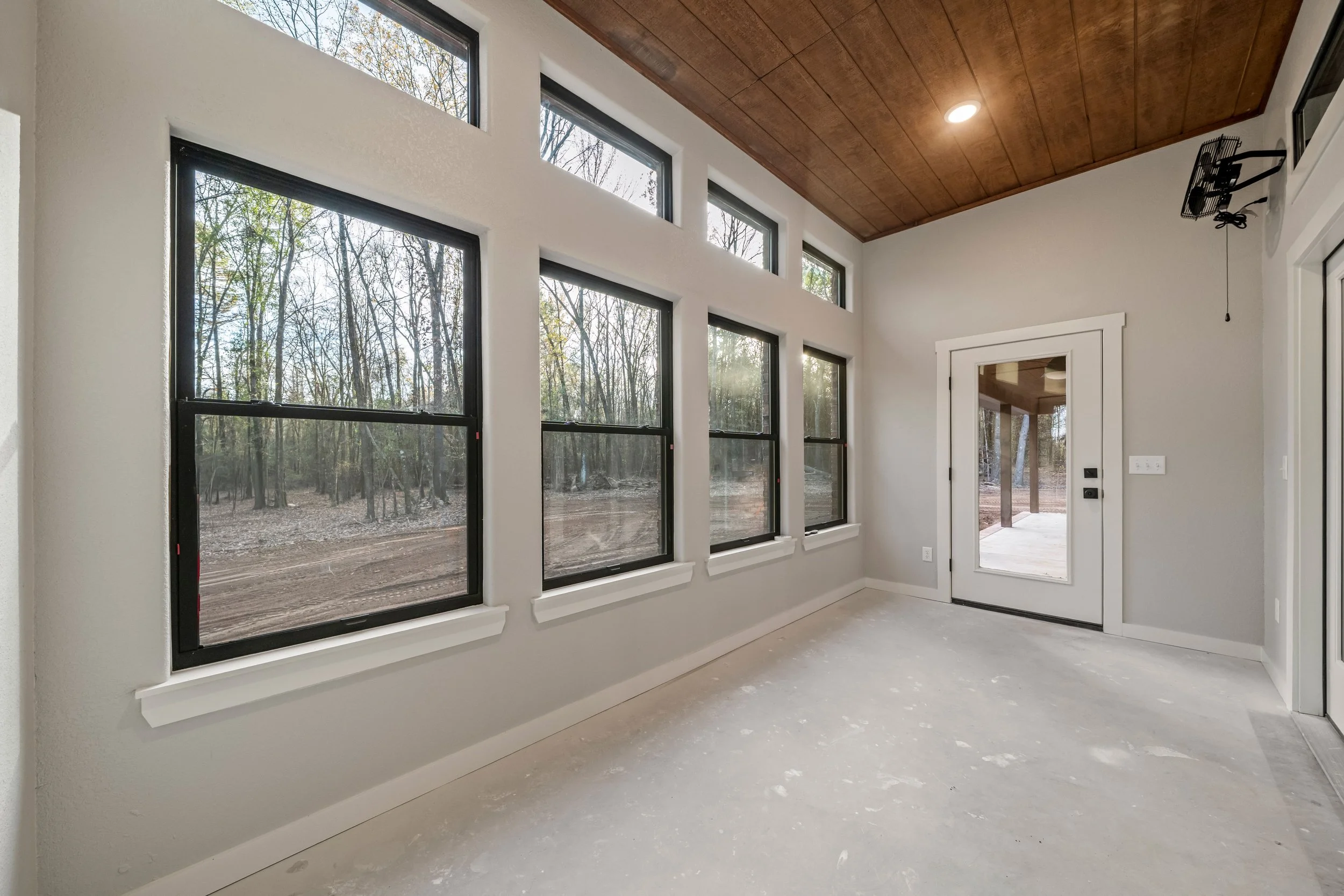 Empty room with multiple large windows facing a wooded outdoor area, a glass door leading outside, and a wood-paneled ceiling.