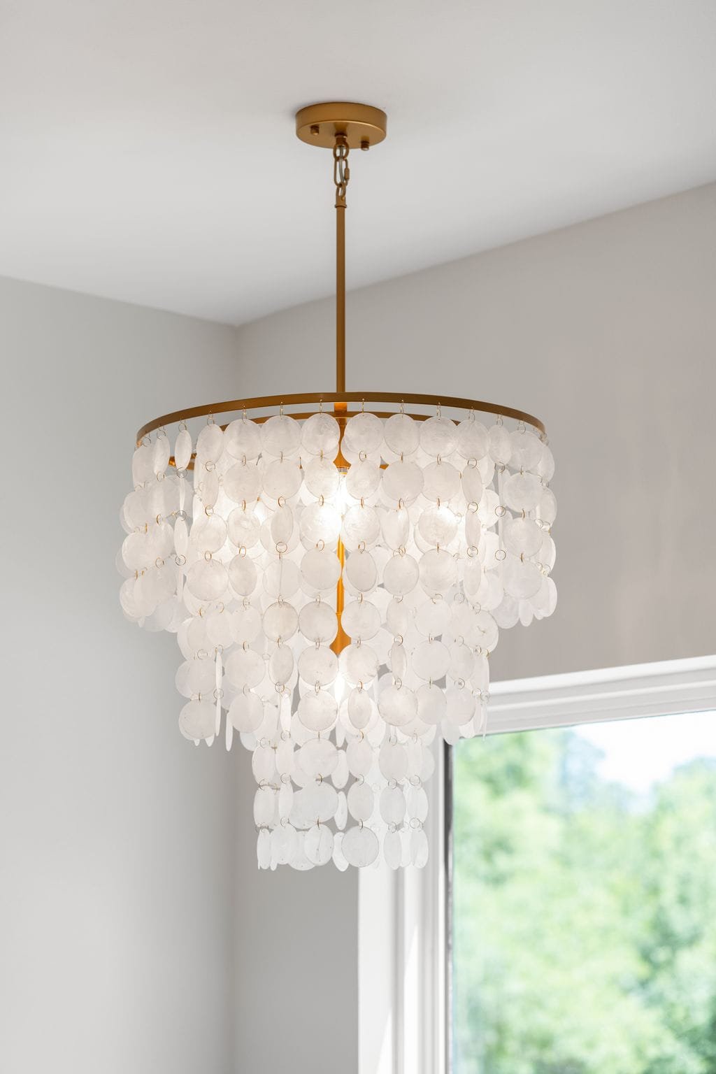 A white chandelier with circular shell-like ornaments hanging from a circular gold frame, mounted on a white ceiling near a window with green trees outside.