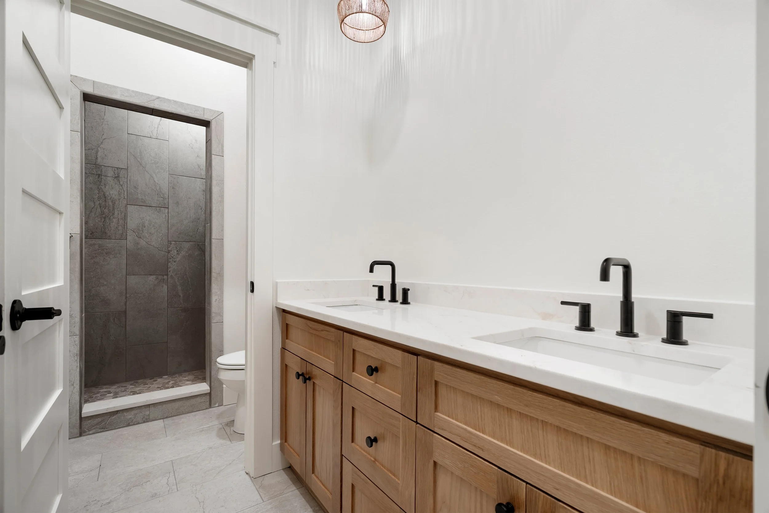 Modern bathroom with double sink vanity, marble countertop, black fixtures, wooden cabinet, and a separate shower area with gray tile walls.