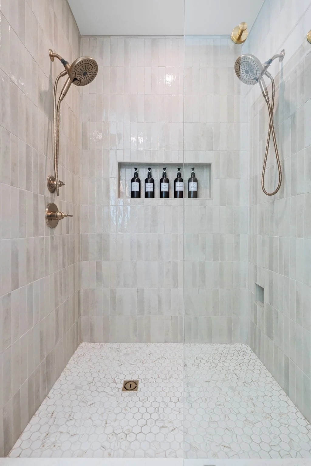 Walk-in shower with dual showerheads, vertical tile surround, recessed niche shelving, and hexagon tile flooring.