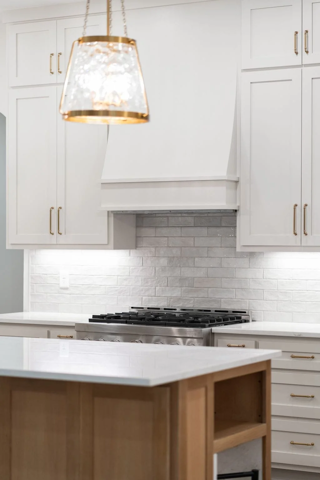 A modern kitchen with white cabinets, a white brick backsplash, a stainless steel gas stovetop, and an island with a white countertop and wooden base. A stylish gold and glass pendant light hangs from the ceiling.