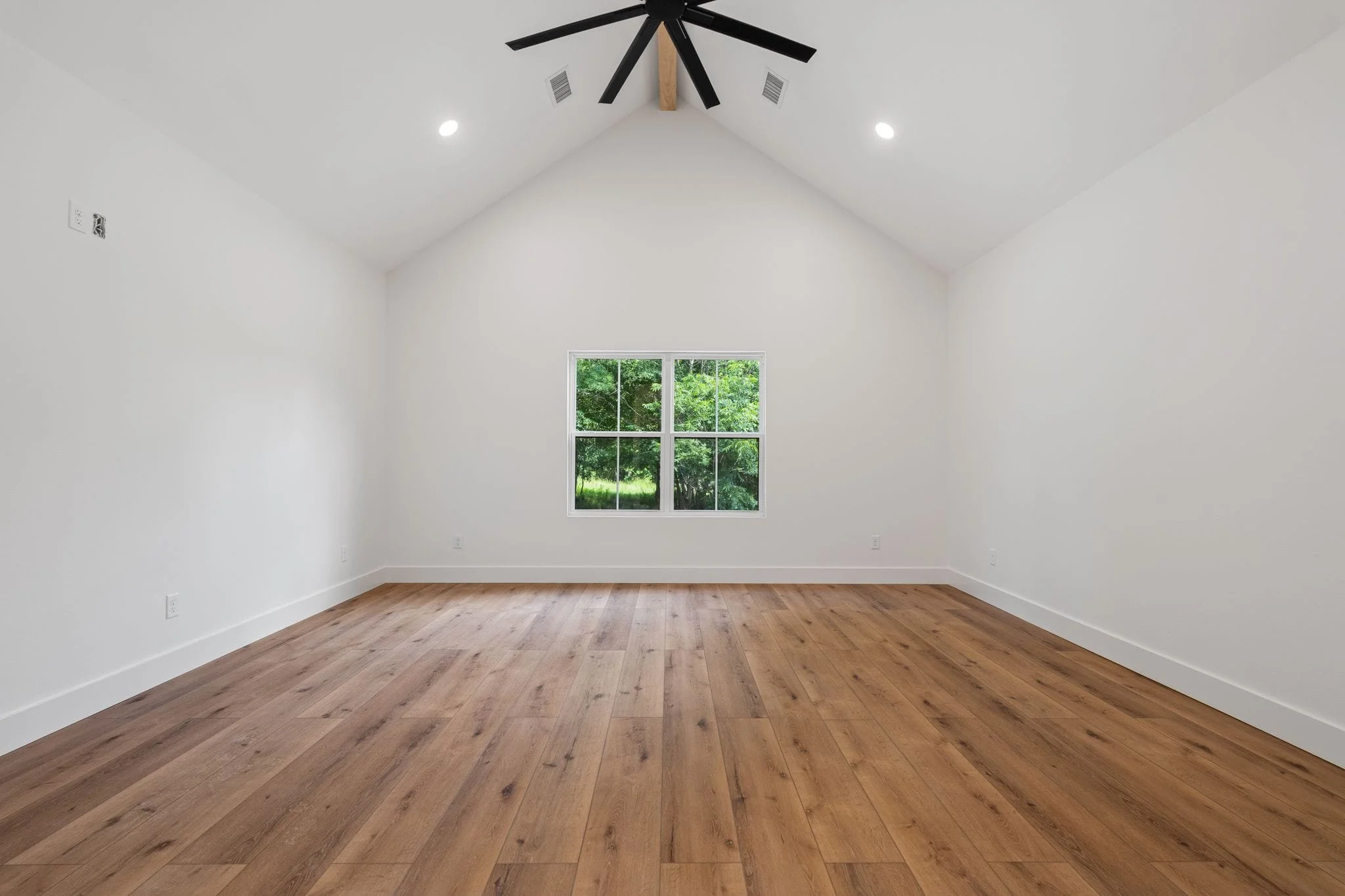 Empty room with white walls, wooden floor, a large window, and a ceiling fan with black blades.