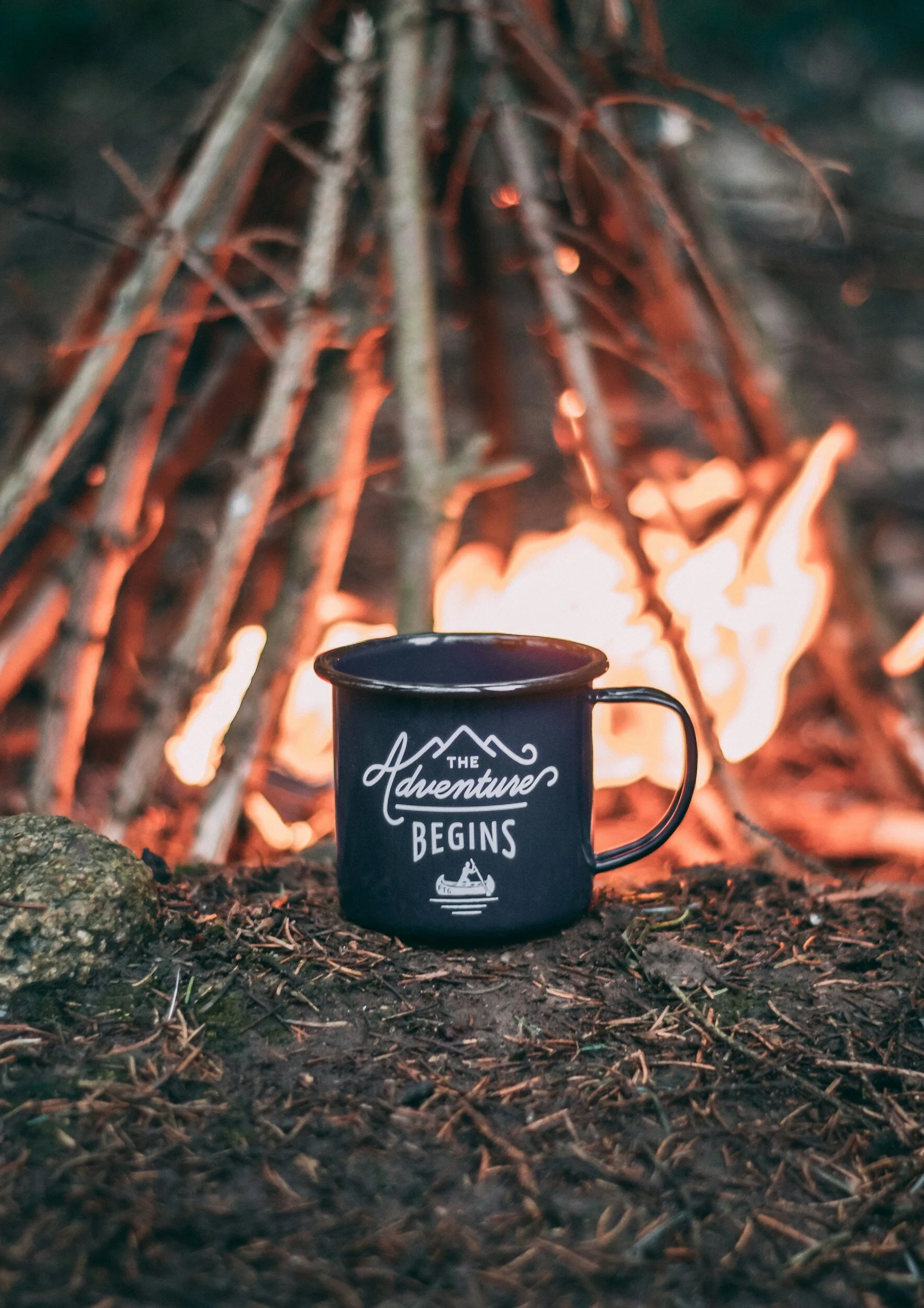 A black enamel mug with the text "The Adventure Begins" stands on the ground in front of a lit campfire, surrounded by sticks and forest floor debris.