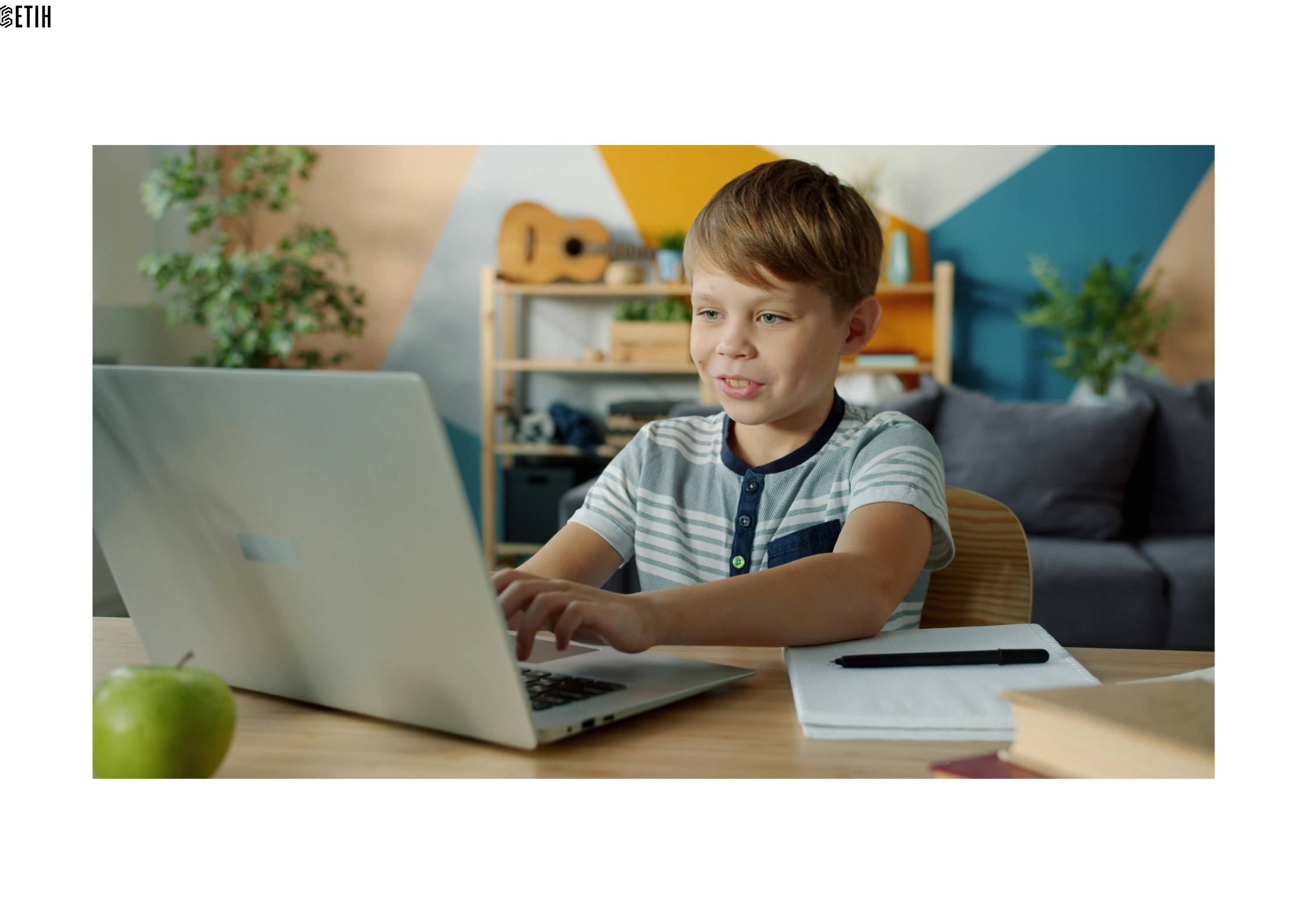 Pictured is a boy working on his laptop with a notebook and pen at his side on the desk