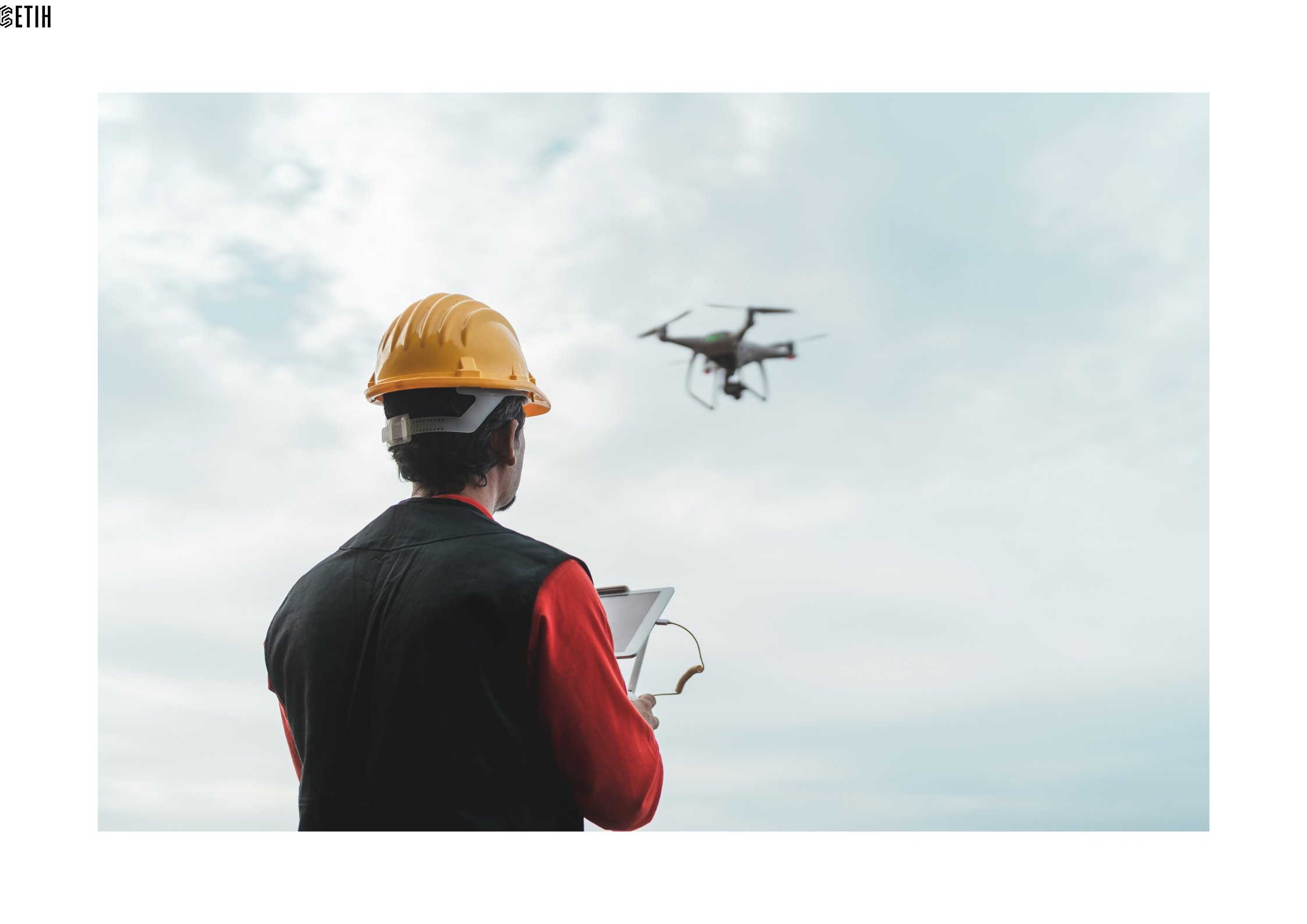 Pictured is a man facing away from us and wearing a yellow hard hat, controlling a drone flying in the distance