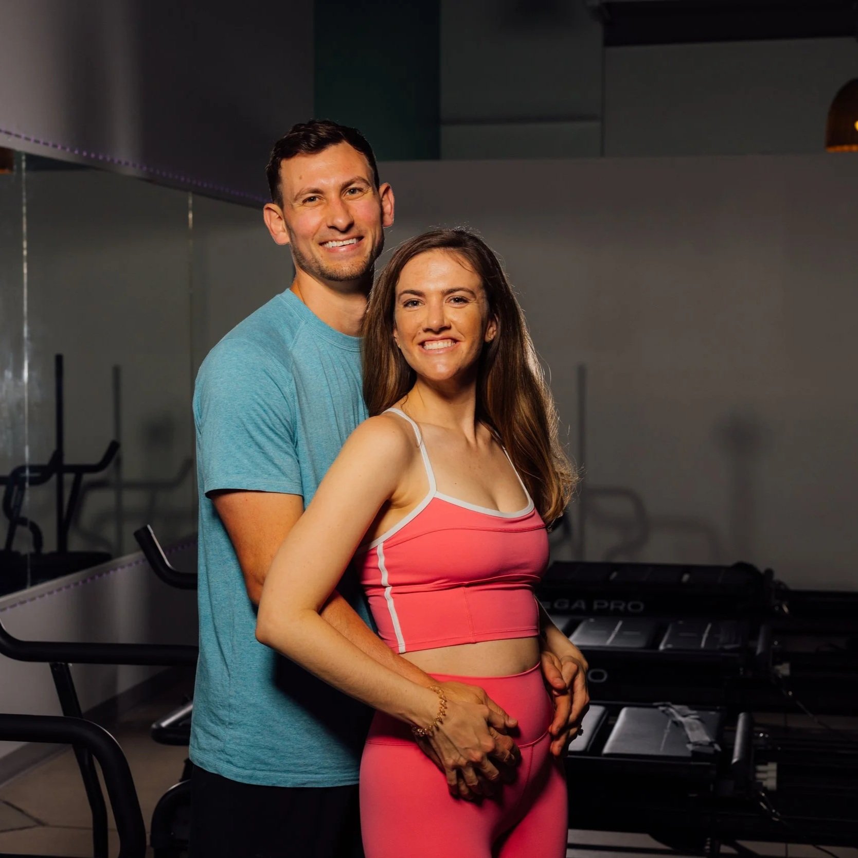 A young man and woman pose together in a gym. The man is standing behind the woman, both smiling at the camera. The woman is wearing a pink sports bra and matching leggings, and the man is wearing a blue t-shirt. The gym equipment is visible in the background.
