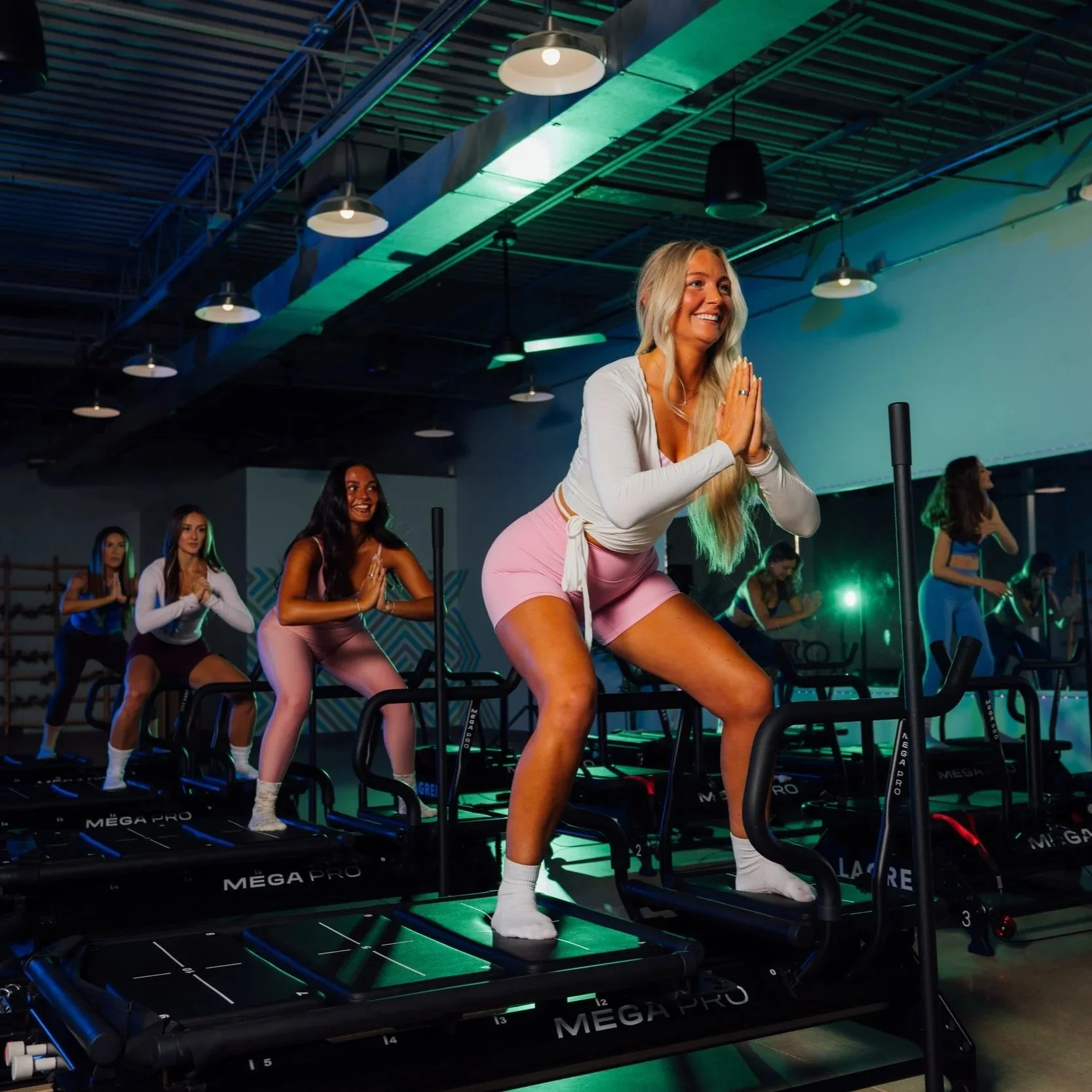 Group of women doing exercise on treadmills in a gym, with a woman in front leading a workout class, smiling, wearing pink shorts and a white top.