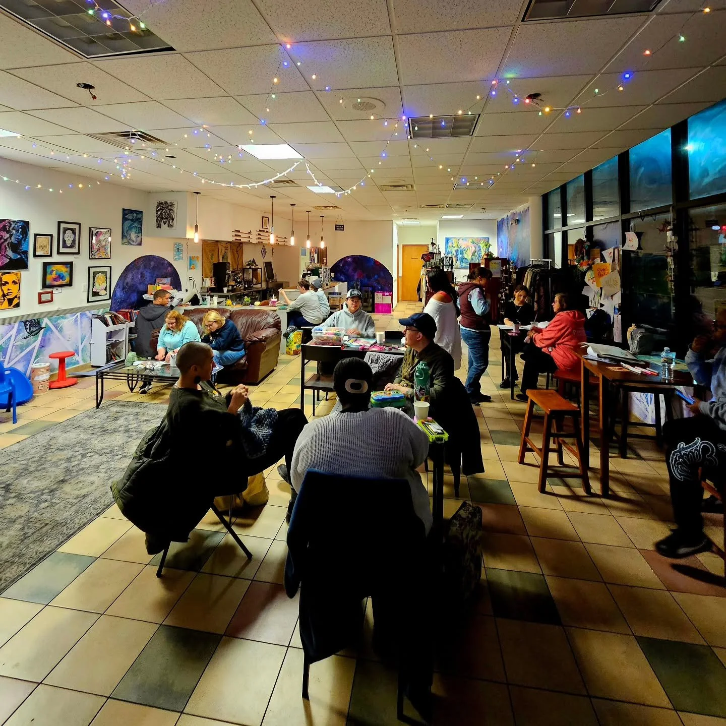 People gathered in a well-lit art gallery or studio with colorful artwork on the walls, tables with art supplies, and string lights hanging from the ceiling.