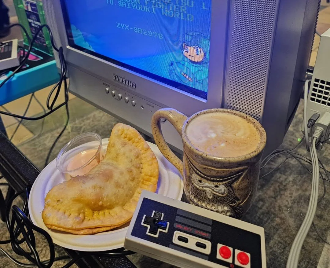 A plate with fried empanadas and a small container of dipping sauce, a large coffee mug with latte art, a Nintendo controller, a Sony TV showing a Sonic the Hedgehog game, and various electronic cables.