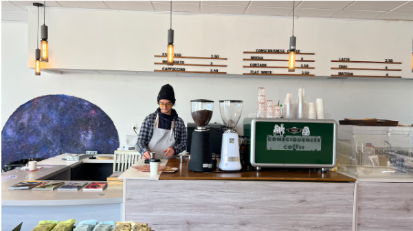 Barista preparing coffee at a coffee shop counter with coffee grinders, cups, and a green espresso machine.