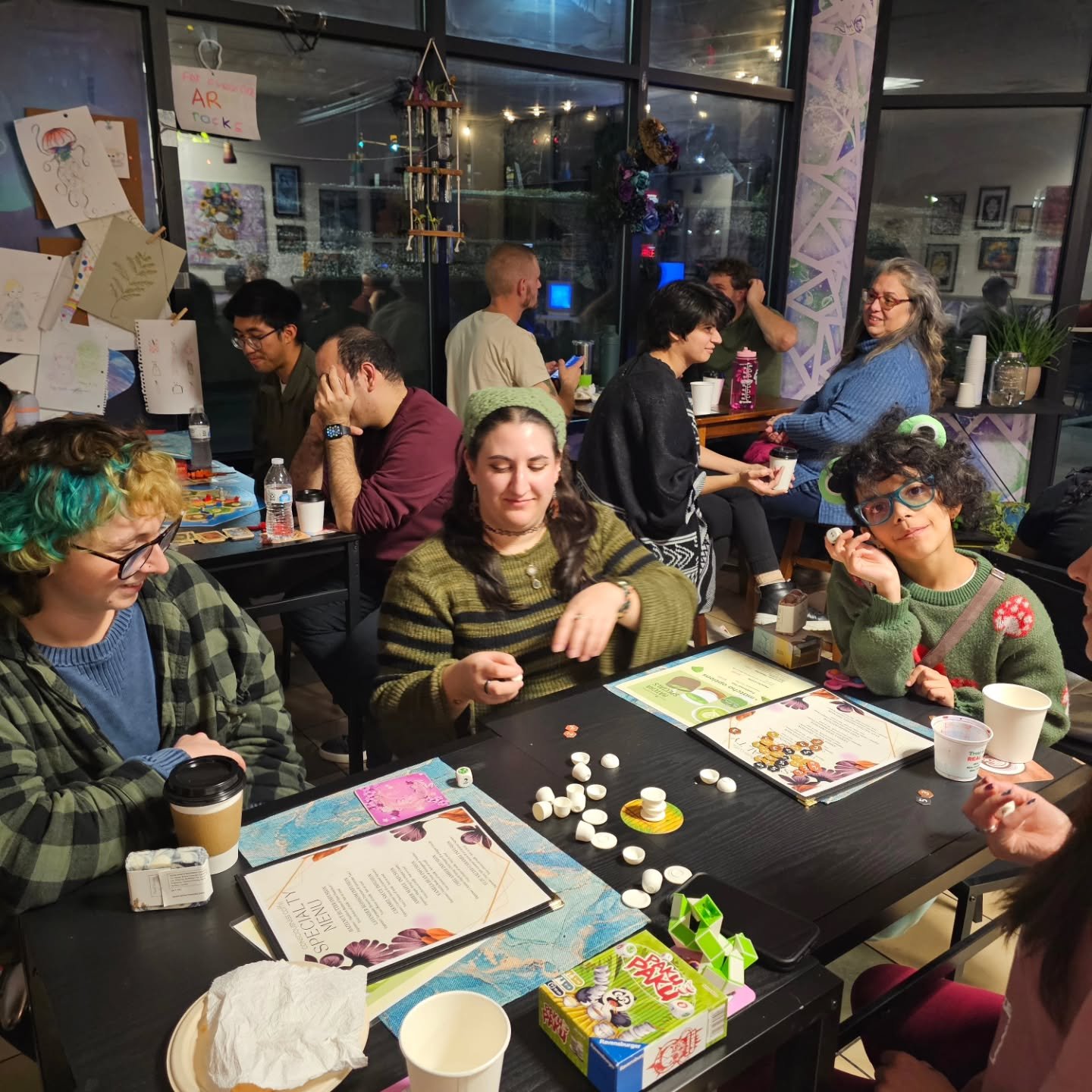 A group of people playing a board game at a table in a cozy cafe or restaurant with artwork on the walls and decorative items