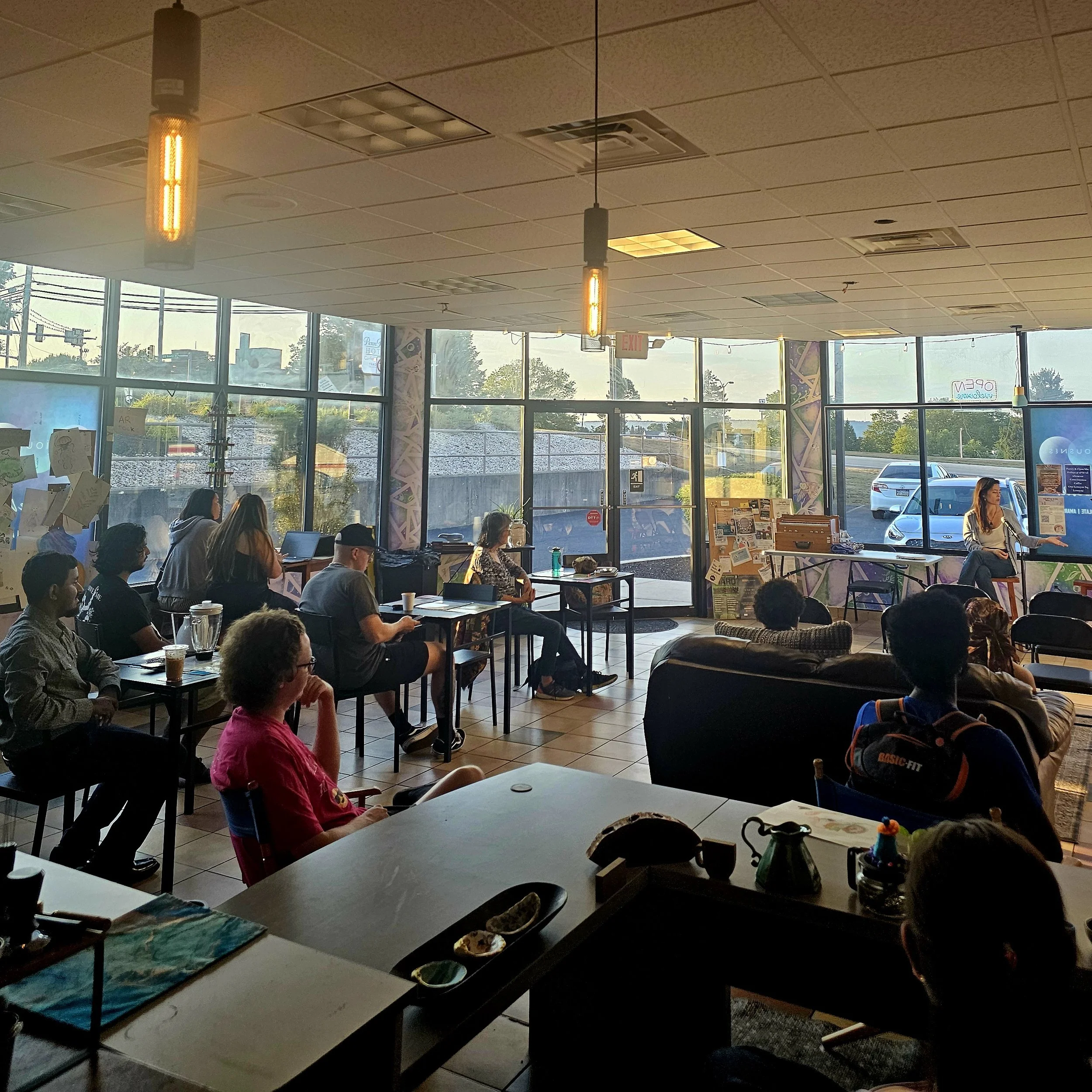 A woman is giving a presentation in a brightly lit coffee shop or café, sitting in front of a large screen with an audience of diverse people seated at tables and couches, some taking notes and others listening attentively.