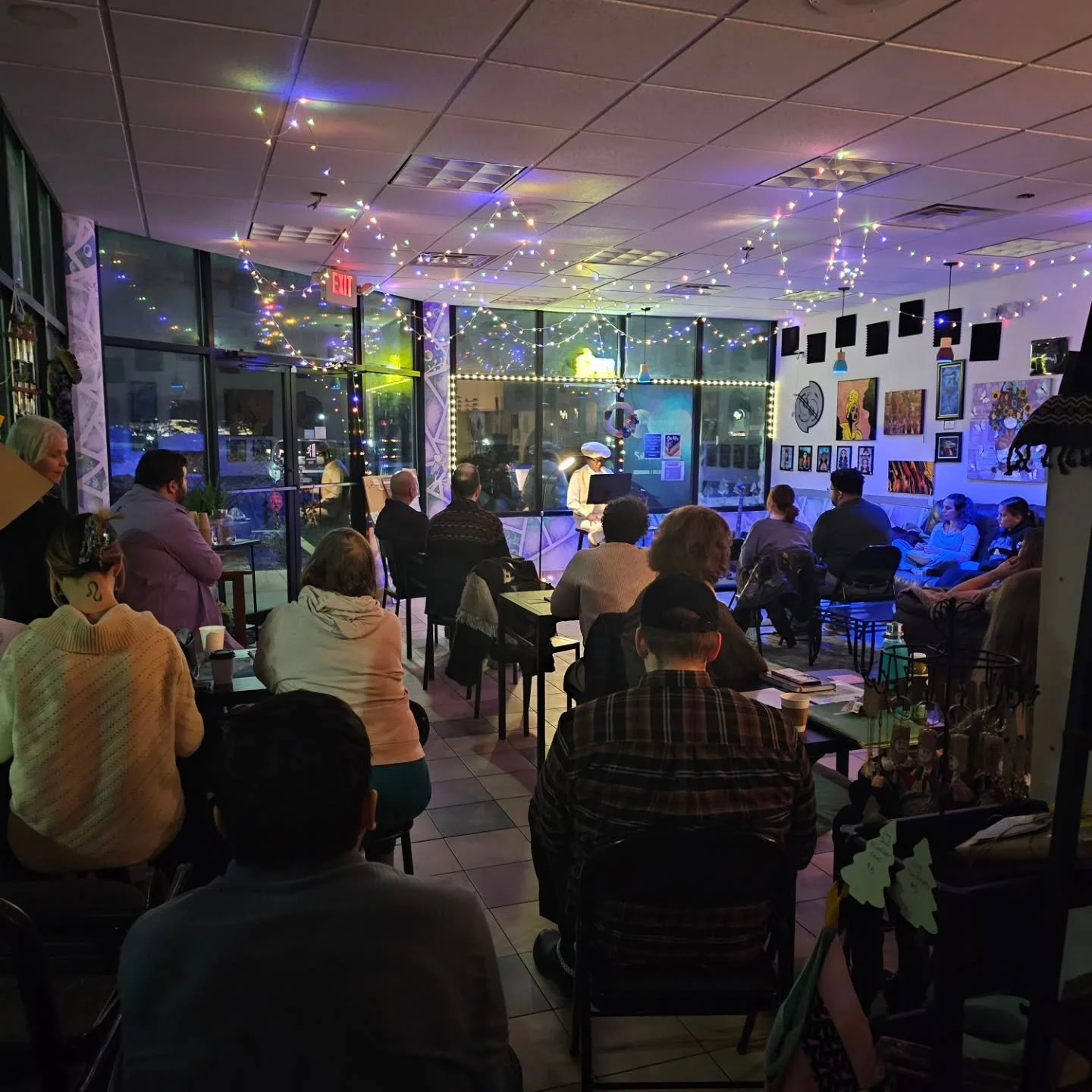 People gathered in a cozy art gallery or cafe with string lights and colorful artwork on the walls, listening to a person in a cowboy hat performing or speaking at the front.