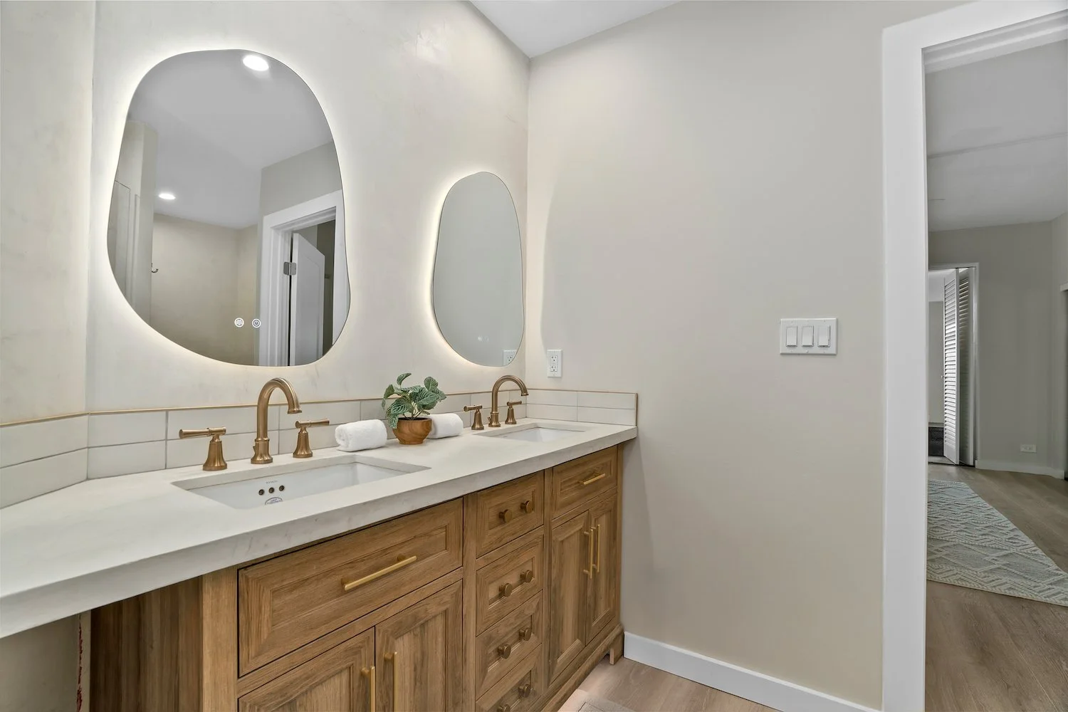 Bathroom vanity with double sinks, gold fixtures, two oval mirrors, a potted plant, and a beige wall in a modern bathroom.