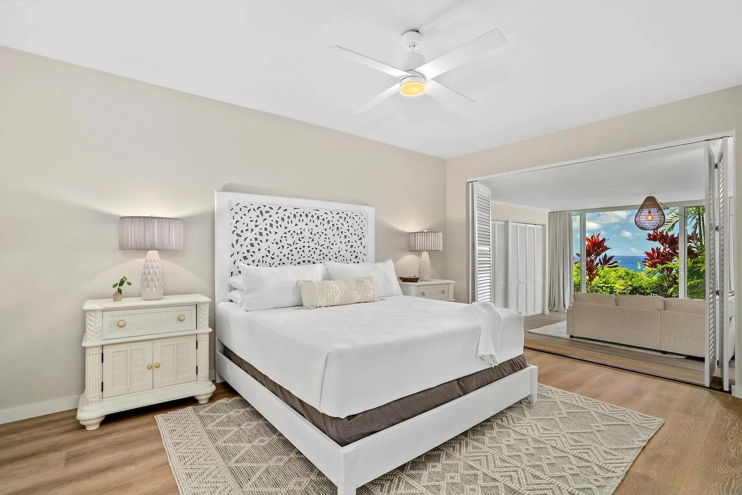 A bedroom with a white bed, nightstands, lamps, and a view of tropical garden and ocean through a large sliding door.