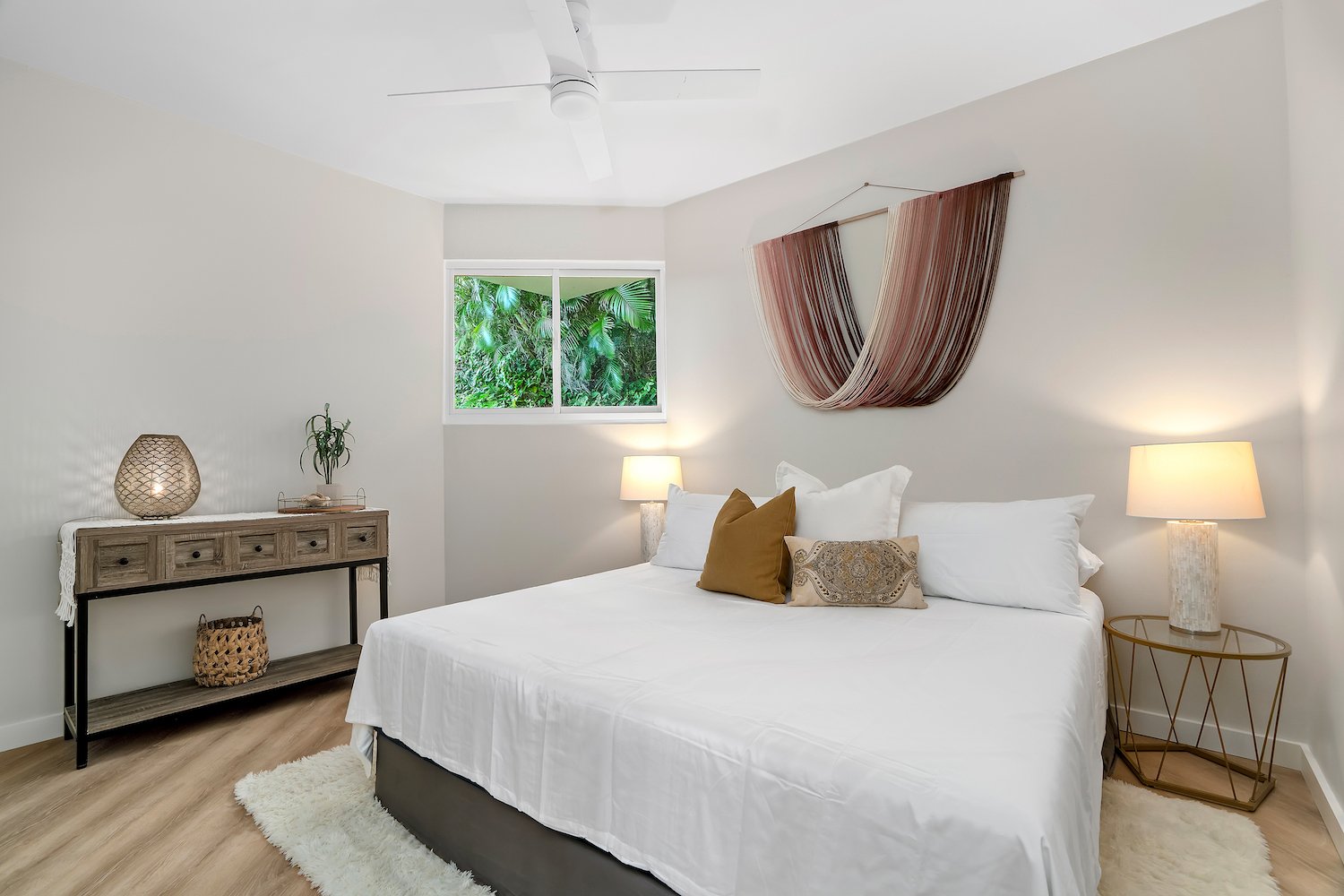 A minimalist bedroom with a large bed covered in white bedding, two bedside lamps, a window with green foliage outside, and a wall hanging above the bed.